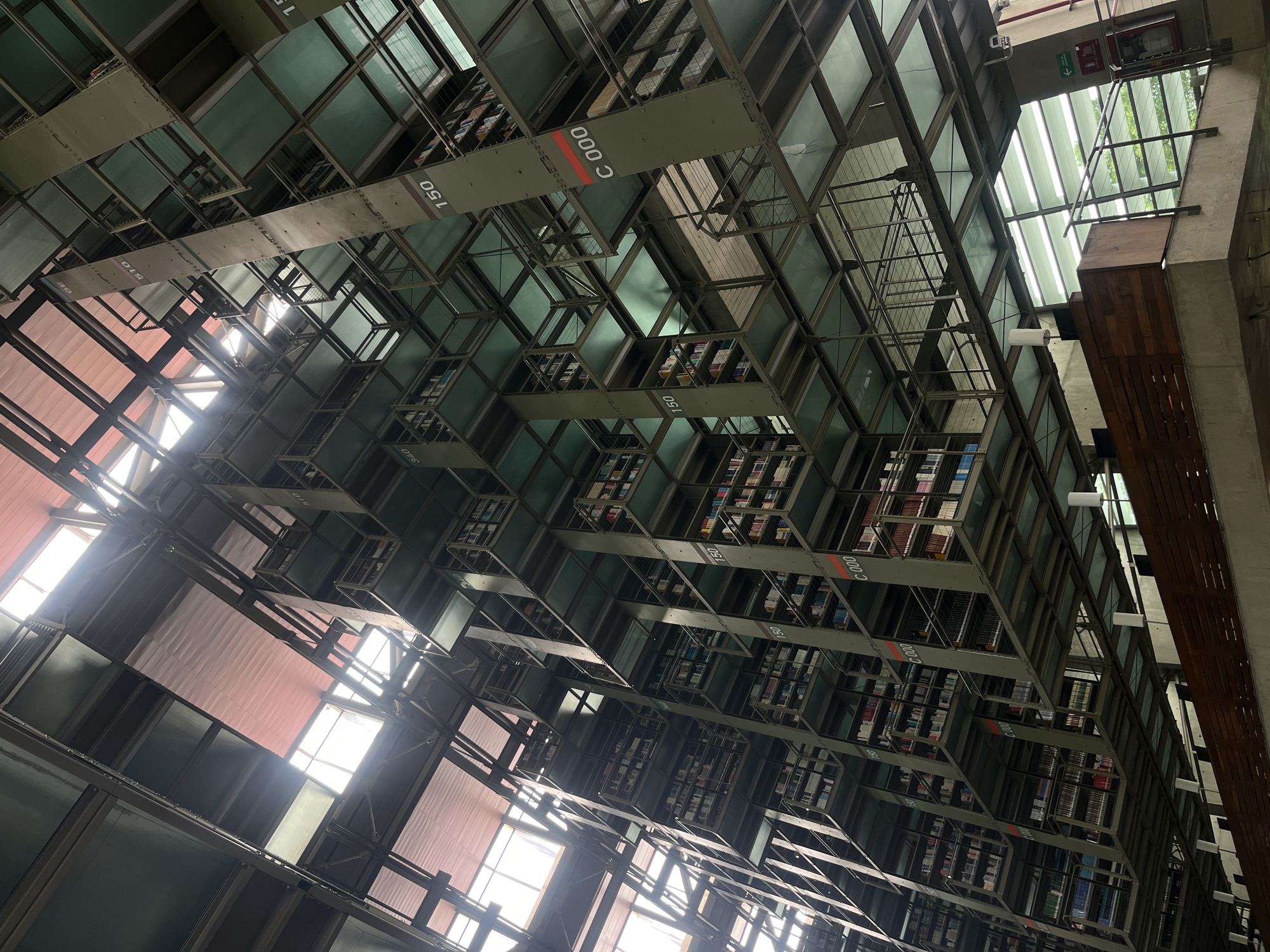 The image shows an interior view of a large, multi-story library with a distinctive architectural design. The bookshelves are arranged in a complex, grid-like structure that extends upwards, creating a visually striking effect. The shelves are made of metal and glass, with many of them filled with books. Sunlight streams in from large windows, illuminating some sections of the library and casting shadows on others, suggesting it is daytime. The overall impression is one of a modern and vast repository of knowledge.
Visible text includes labels on the shelves, such as "C 000" and "150," which likely indicate sections or call numbers. The architecture suggests a public building, possibly a university library or a major city library. The location cues, such as the sunlight and the type of interior construction, are consistent with a warm climate. There are no people visible in the image. The scene evokes a sense of order, scale, and intellectual pursuit.