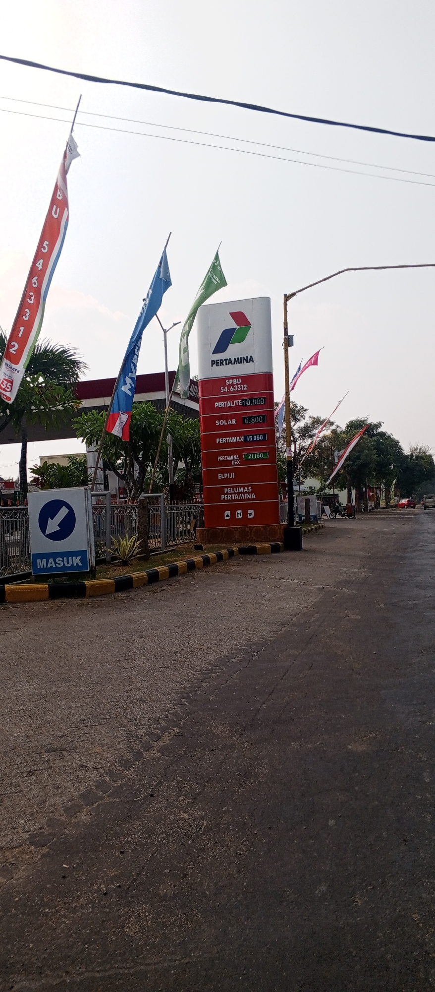 This is a medium shot, eye-level, outdoor, daylight photograph of a Pertamina gas station in Maospati, Indonesia. The gas station is identified by its prominent red and white sign featuring the Pertamina logo and fuel prices. Flags with the Pertamina logo and other designs are flying in front of the station. The road in front of the gas station is asphalt with yellow and black striped curb markings. A blue and white sign with a left-pointing arrow and the word "MASUK" (Indonesian for "ENTER") is visible to the left of the frame. The sky is bright and overcast. Trees and a red-roofed building are visible behind the gas station. A motorcycle and a red car are parked in the background, and there are also other Pertamina flags visible further down the road. The scene appears to be a typical street view of a gas station in a town.
