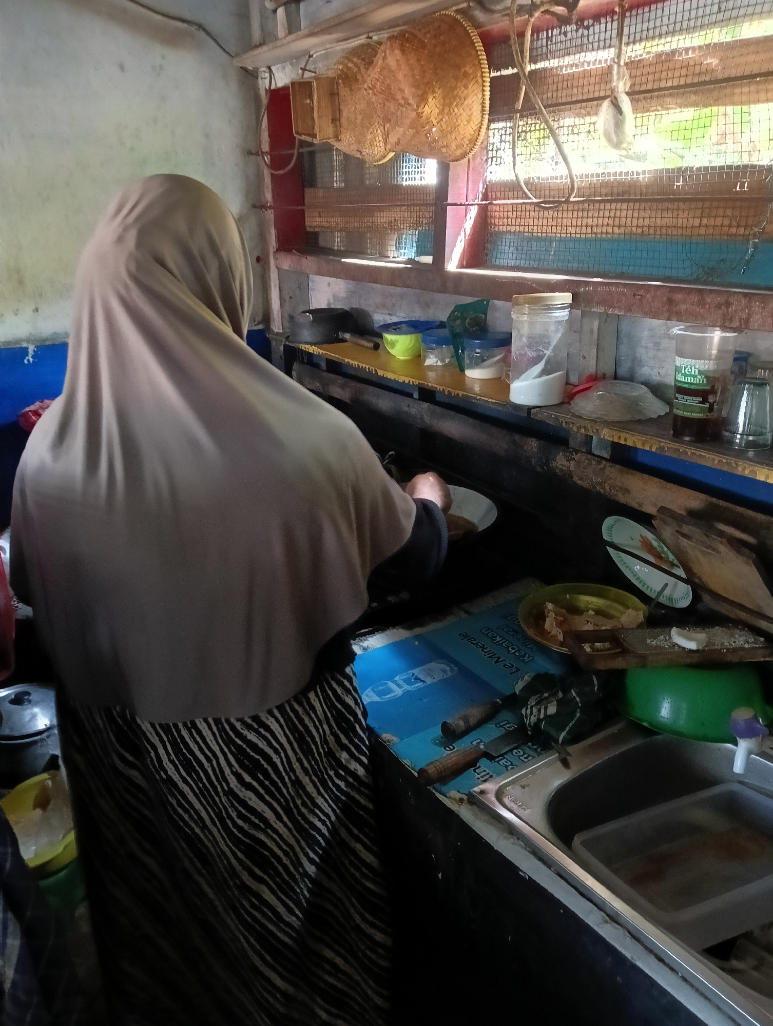A person wearing a beige hijab and a black and white striped skirt is standing at a kitchen counter, facing away from the camera. They are holding a plate in their left hand and appear to be preparing food. The kitchen counter is cluttered with various items, including plates, containers of food, a sink, and utensils. Sunlight streams in from a window behind the counter, illuminating the scene. The kitchen appears to be in a modest home or small business in Manggari, Indonesia. The walls are painted a light color, and the counter is made of wood. There are shelves above the counter holding more kitchen supplies. The overall impression is one of a functional, working kitchen.