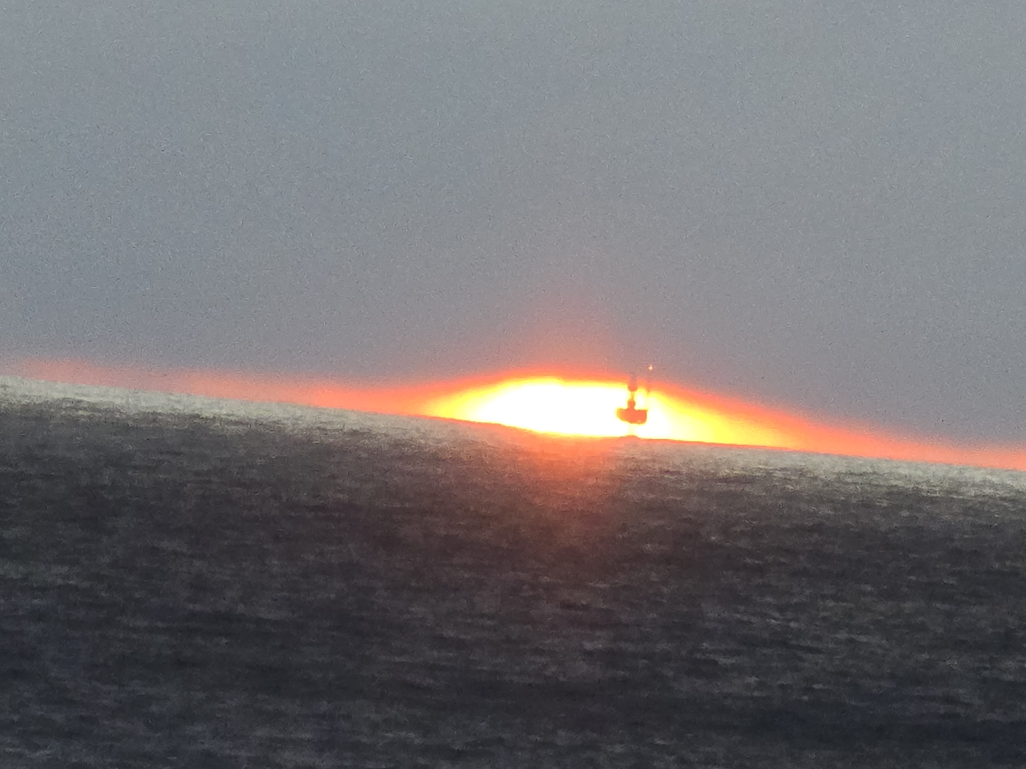 The image depicts a horizon line, likely at sea, with the sun setting or rising. The sun is a bright, diffuse glow, partially obscured by the horizon, creating a band of orange and red light across the sky. Silhouetted against the sun is a small object, possibly a boat or buoy. The lower portion of the image shows what appears to be the dark, textured surface of water. The sky is a uniform grey, indicating a cloudy or overcast condition. The location is Unknown, Unknown.