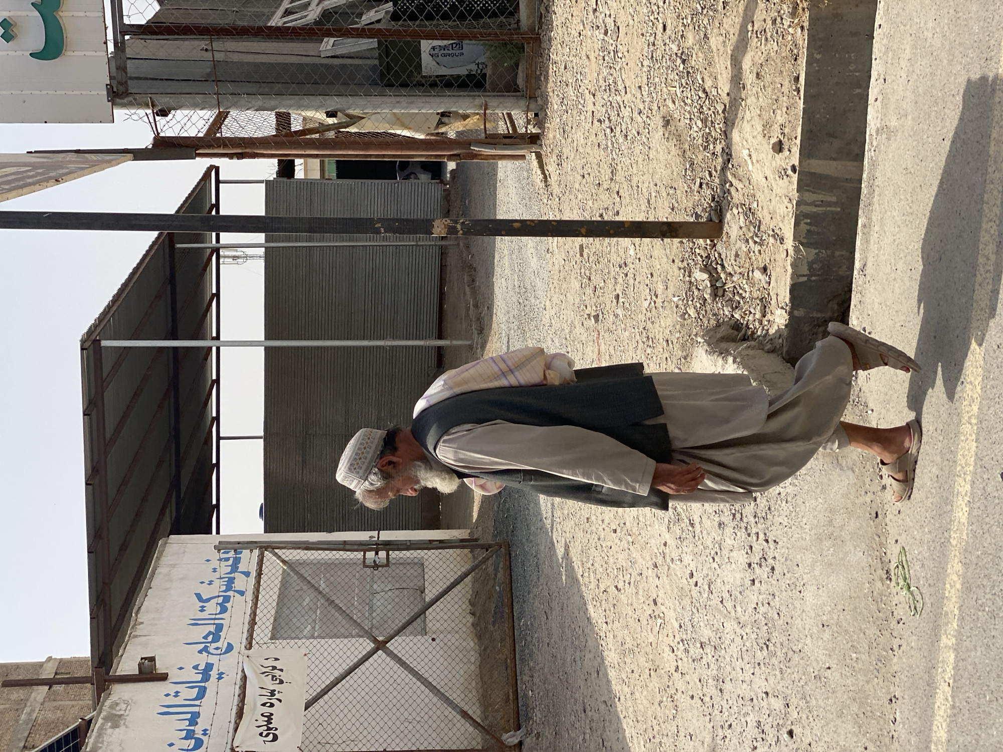 The image depicts an outdoor scene in the city of Kandahar, Afghanistan. A man, likely an elder, with a grey beard and wearing a traditional white patterned cap, a light-colored shirt, a dark vest, and loose-fitting trousers, is walking from left to right. He carries a folded cloth over his right shoulder. The man is walking on a paved surface adjacent to a dirt and gravel area. A thin, green cord lies on the paved surface in the lower left of the frame.
In the background, there is a white building with blue Arabic script painted on its facade. A chain-link fence separates the paved area from the building. Beyond the fence, there are corrugated metal structures and a covered open-air area with a metal roof supported by posts. To the right, a metal fence with wooden planks visible behind it encloses further structures. The sky is overcast and appears pale. The overall setting suggests a commercial or industrial area.
