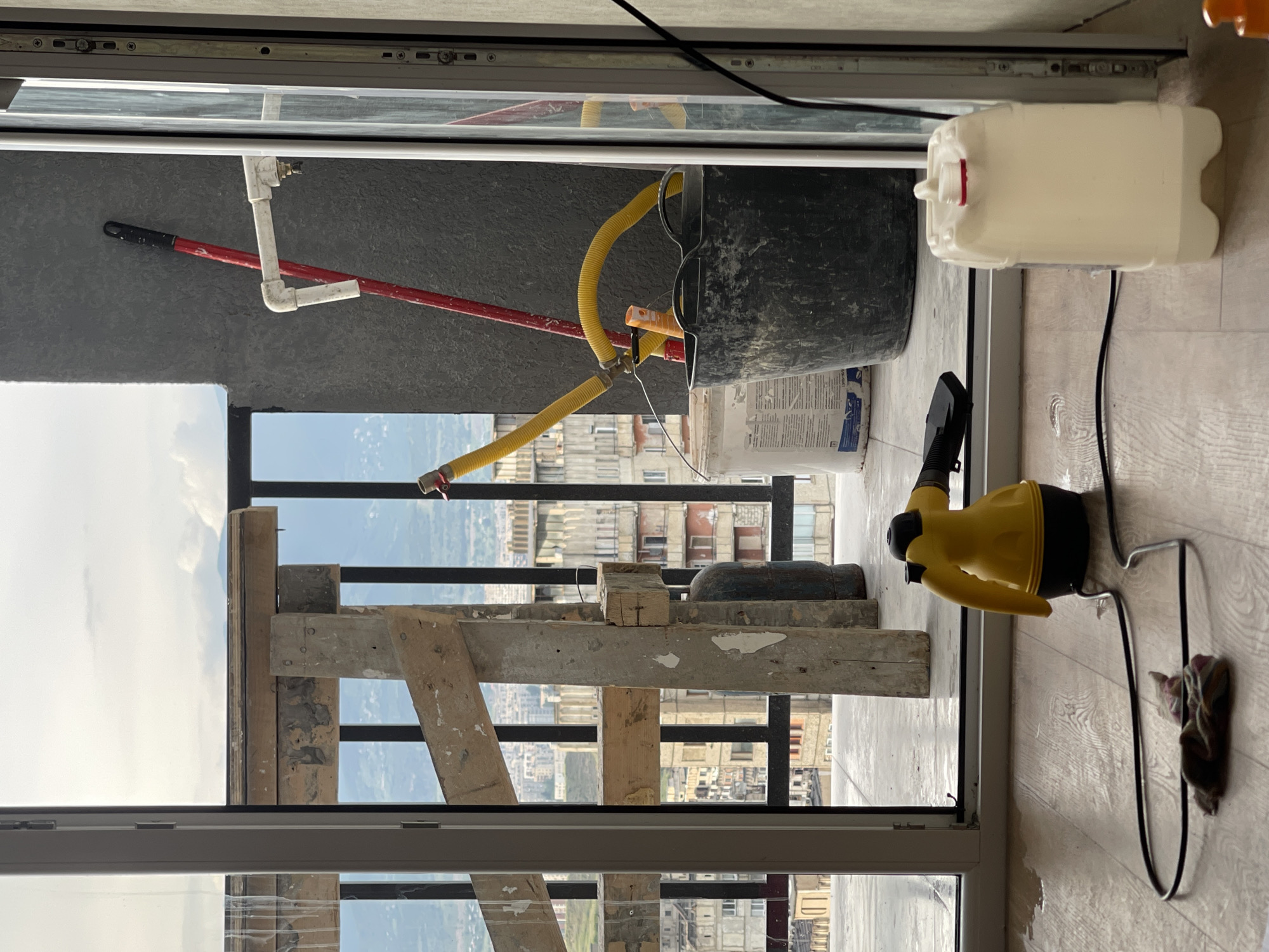 The image depicts a balcony area of a building under construction or renovation, overlooking a cityscape that is consistent with Tbilisi, Georgia. In the foreground, a yellow steam cleaner with a black hose is placed on a light wooden floor, near a small pile of cloth items and a white plastic container filled with liquid. The floor appears wet in places, suggesting recent cleaning or a spill.
Further back, on the balcony itself, a large black bucket is visible, containing yellow hoses connected to an orange object. A red broom leans against the textured gray wall, near a white pipe fixture. Various construction materials are present, including wooden planks forming scaffolding, concrete pillars, and large bags or barrels.
The background reveals a dense urban environment with multi-story buildings, characteristic of Tbilisi's architecture, nestled within a valley. The sky is overcast with clouds. The scene suggests a pause in construction or cleaning activities, with tools and materials laid out. There are no people visible in the image.
