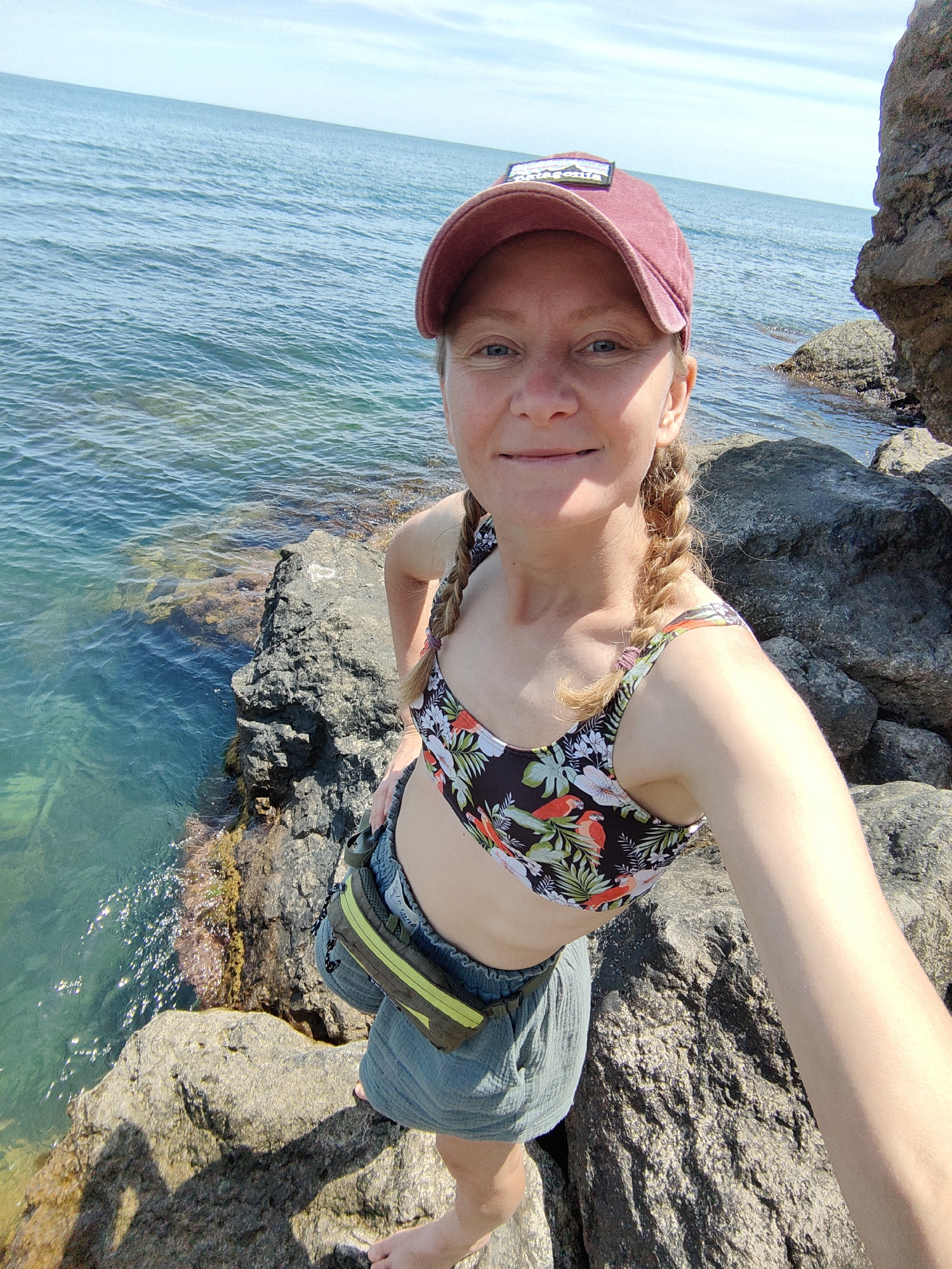 A woman is taking a selfie on a rocky coastline. She is wearing a red baseball cap, a floral print bikini top, and grey shorts. The ocean is visible behind her, with a clear, blue-green color and gentle waves. There are large rocks surrounding her, some of which are partially submerged in the water. The sky is blue with a few scattered clouds. The location is identified as Utios, Ukraine.