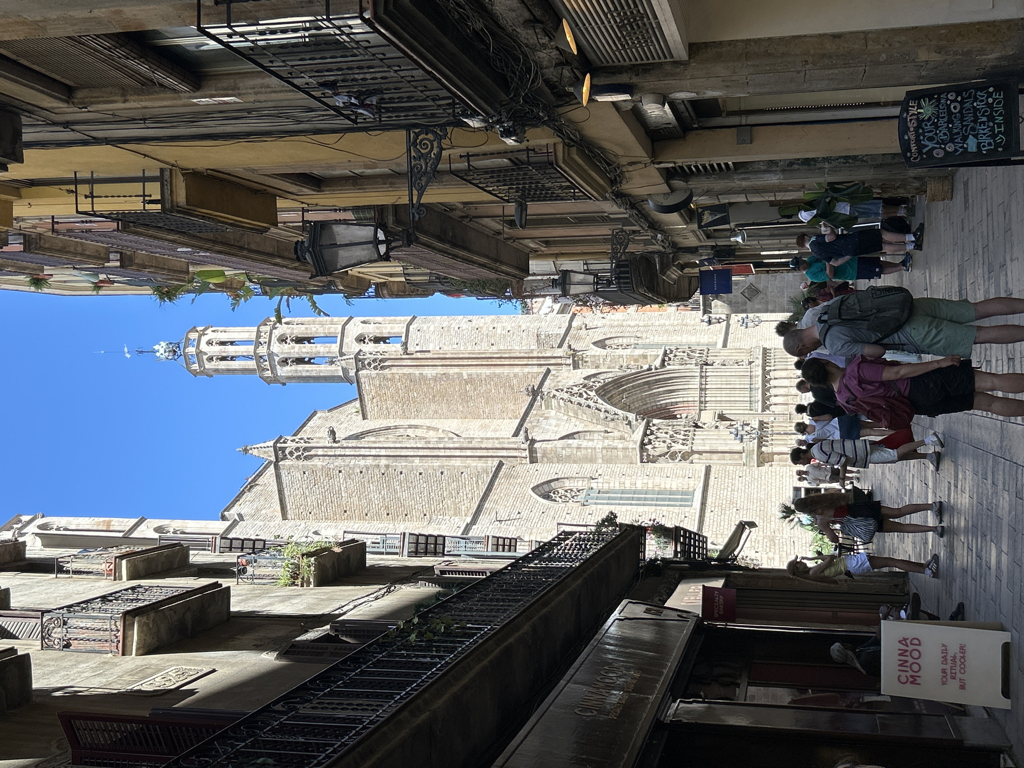 The image captures a view down a narrow street in Barcelona, Spain, with a grand Gothic cathedral dominating the background. The cathedral, built of light-colored stone, features intricate architectural details, including pointed arches, flying buttresses, and a prominent bell tower topped with a spire. On either side of the street are multi-story buildings with balconies and shutters. The building on the left appears older, with stone facades and dark balconies. The building on the right is painted a yellow hue and features several balconies, some adorned with plants, and external lighting fixtures.
A group of people are walking and standing in the street, some appearing to be tourists. In the foreground, on the left, there is a sandwich board sign with the text "CINNAMOOD YOUR DAILY RITUAL, BUT COOLER!". Another sign in the lower right corner reads "COMFORT STYLE YOUR BARCELONA WALKING SANDALS BIRKENSTOCK INSIDE". The street is paved with cobblestones. The sky is a clear, bright blue. The sun casts distinct shadows, indicating it is likely midday.