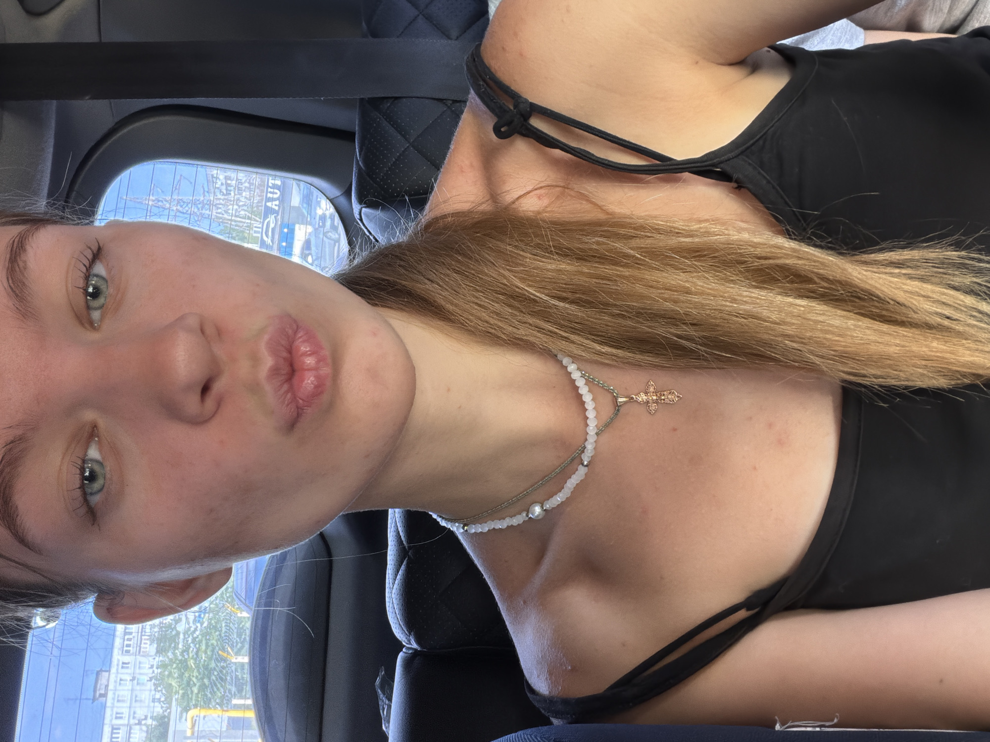A blonde woman with green eyes and fair skin is captured in a close-up shot, leaning forward with her lips puckered. She is wearing a black tank top and a delicate necklace with a pearl and a small gold cross. Her hair is long and blonde, pulled to the side. In the background, visible through the car window, are buildings and what appears to be a street scene, suggesting she is inside a vehicle in an urban environment. The context provided identifies the location as Rostov-on-Don, Russia. The overall mood is casual and personal, likely a selfie taken while traveling.