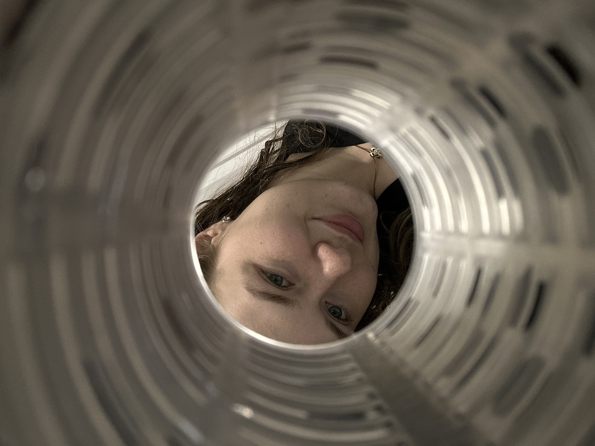 A woman is seen through the opening of a cylindrical metal structure. The focus is on her face, which is tilted towards the camera. She has fair skin, light eyes, and dark hair. She is wearing a necklace and earrings. The interior of the cylinder has a repeating pattern of dark markings along its surface. The image was not taken in Studenets, Russia.