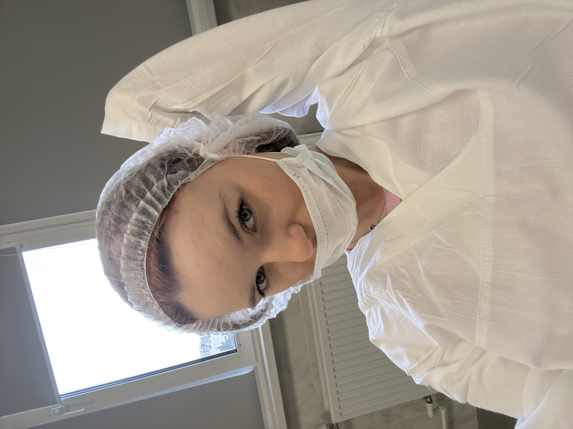A person is wearing a white surgical gown, bouffant cap, and surgical mask. The person is indoors near a window. The location appears to be in Saint Petersburg, Russia. The person is looking down at the viewer. A radiator is on the wall.