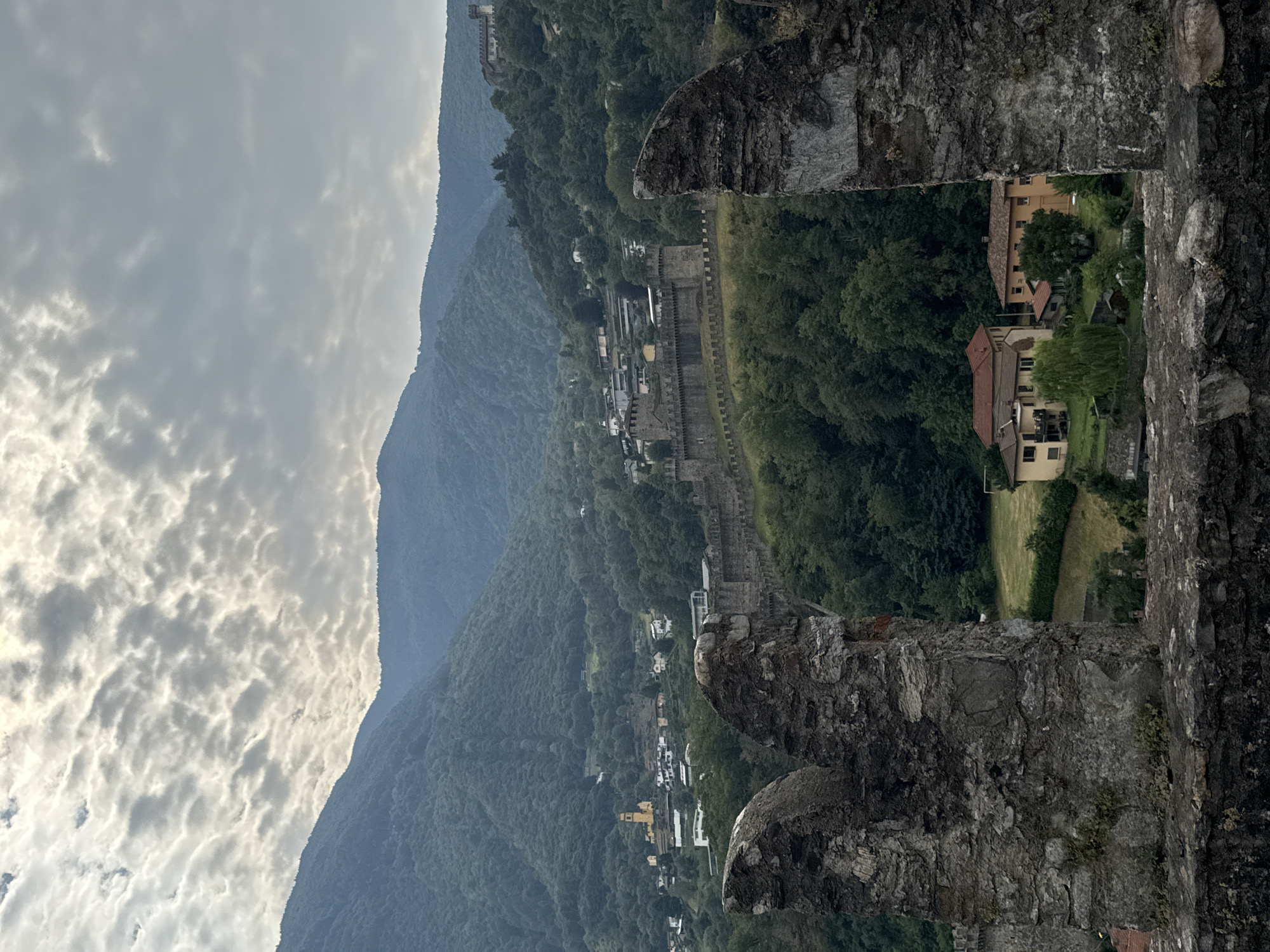 The image captures a view of Castel Grande, one of the three castles of Bellinzona, Switzerland, set against a backdrop of lush green mountains under a cloudy sky. In the foreground, rough-hewn stone battlements suggest the viewer is positioned within a similar ancient structure. Below, a village nestles on the slopes, characterized by terracotta-tiled roofs and yellow-hued buildings. A prominent yellow church spire stands out in the mid-ground. The main castle complex, a long stone wall with crenellations, stretches across the landscape, with additional towers and buildings visible further up the hillside, indicating the fortifications extend across multiple levels. The environment appears serene, with dense foliage covering the surrounding mountains and manicured lawns visible near the houses. No people are discernible in the frame.