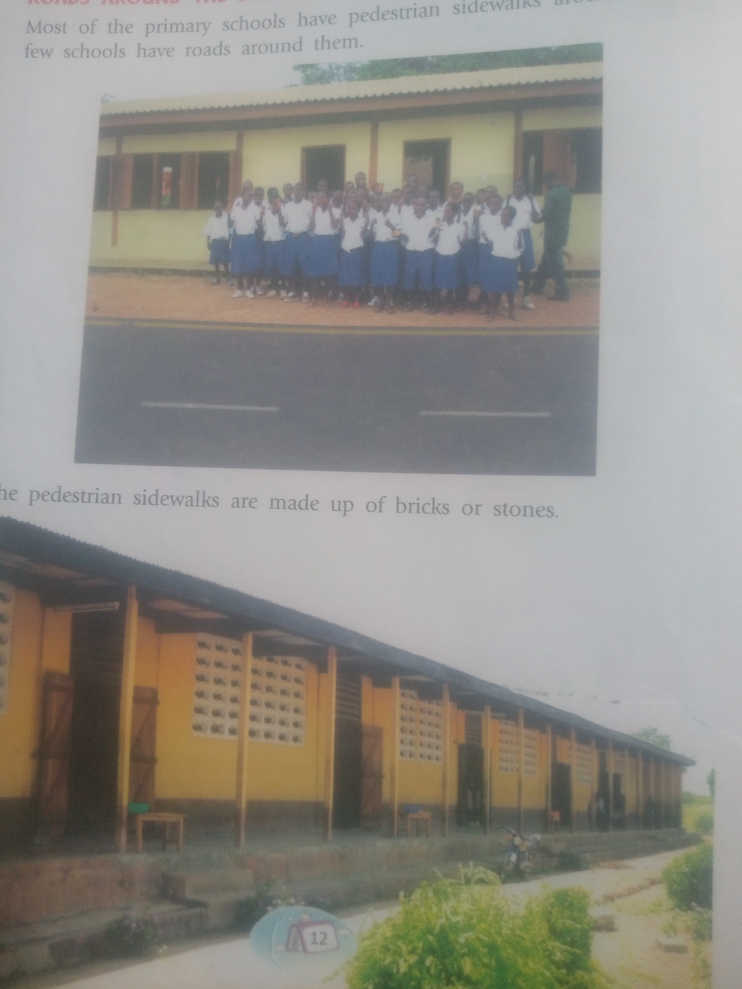 The image contains two photographs of school buildings. The top photograph shows a group of young students in uniform standing in front of a yellow school building. The students are wearing white shirts and blue skirts or shorts. A road is visible in front of the building, with a pedestrian sidewalk. The bottom photograph shows a long, yellow school building with a dark roof. There are several windows with ventilation blocks on the building. A motorcycle is parked in front of the building. The scene appears to be in a rural or semi-urban area. The text below the top photograph states "Most of the primary schools have pedestrian sidewalks and few schools have roads around them." The text below the top photograph continues, "the pedestrian sidewalks are made up of bricks or stones."