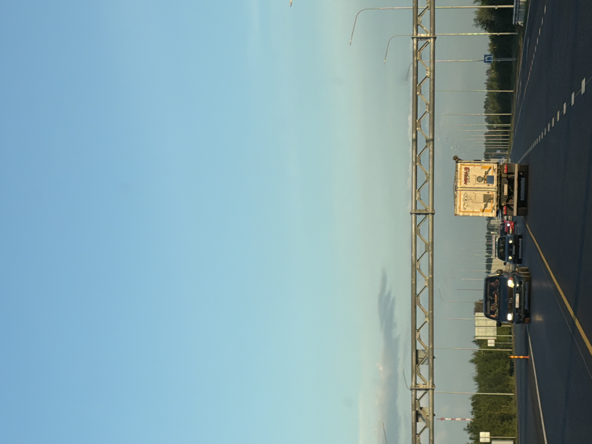 The image depicts a view from a moving vehicle on a multi-lane highway, likely in the vicinity of Kaipy, Russia, given the context. The sky is a clear, pale blue, with some faint clouds visible near the horizon. A large metal overhead gantry, used for traffic signs or lighting, spans the highway. On the right side of the highway, a row of streetlights stretches into the distance. The road surface is asphalt, with clear yellow and white lane markings.
In the foreground, a white truck with a box trailer is traveling in the right lane. The truck appears to be a commercial vehicle, possibly for logistics or delivery, with some markings on its trailer that are partially obscured or difficult to discern. To the left of the truck, a dark-colored van or minivan is also traveling in the right lane, followed by other vehicles. Further down the highway, a black sedan is visible in the left lane. On the left side of the highway, a dense line of green trees forms a backdrop, with a few white road signs placed intermittently. The overall scene suggests daytime traffic on a highway.