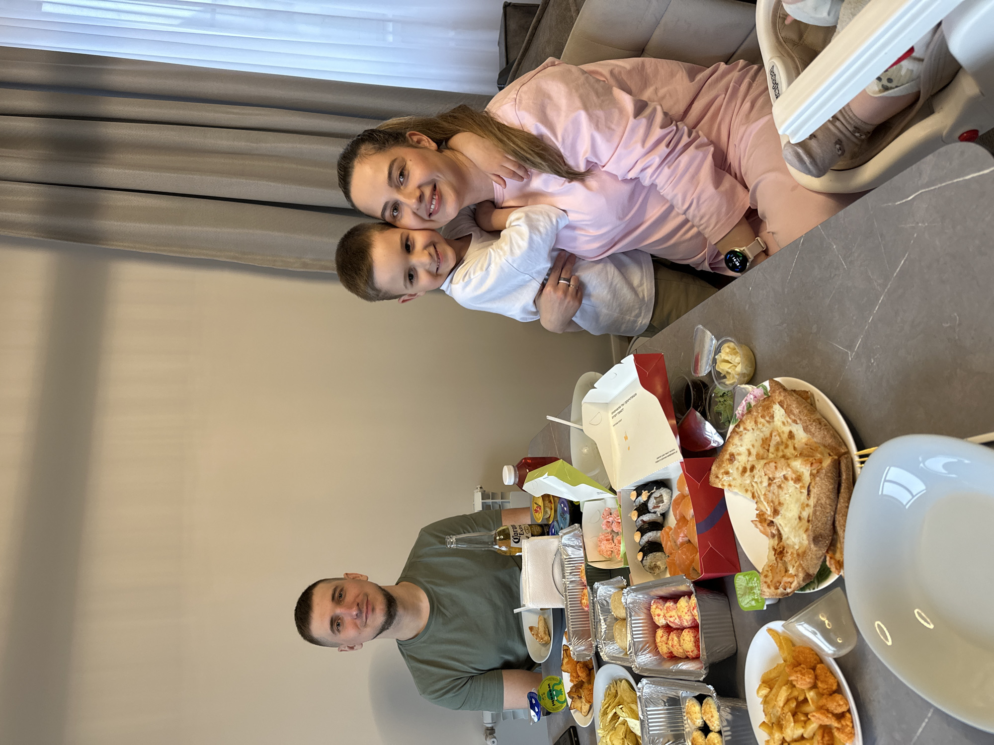 A family gathers at a gray marble table laden with takeout food, including sushi, pizza, and french fries. A man in a green t-shirt stands to the left of the table. A woman in a pink sweatsuit sits at the edge of the table with a young boy, both smiling. A baby in a high chair sits next to the woman. Various beverages, including Corona beer and juice, are visible. The setting appears to be a home dining area with light gray walls and a gray curtain. The city is Chaikovsky, Russia.