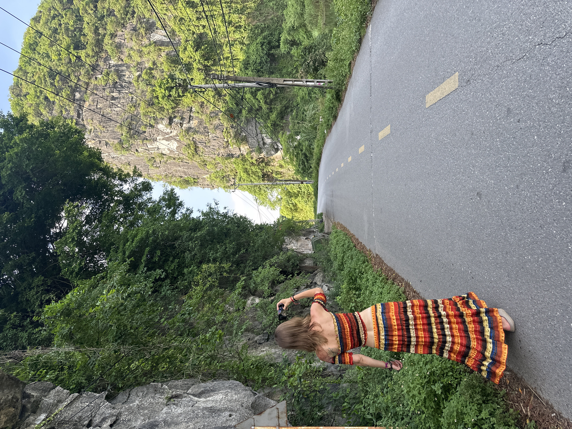 A woman is standing on an asphalt road, facing away from the camera, and taking a photo with her phone. She is wearing a multi-colored, striped off-the-shoulder top and a matching maxi skirt. The road has yellow dashed lines. To her left, a steep, rocky cliff covered in lush green foliage rises up. To her right, the road curves away, bordered by more greenery and a body of water. In the background, another large, imposing karst mountain is visible, also covered in vegetation, with power lines running across it. The sky is clear and blue. The scene appears to be in Nam Hoa Lư Ward, Vietnam, characterized by its dramatic limestone karst landscapes.