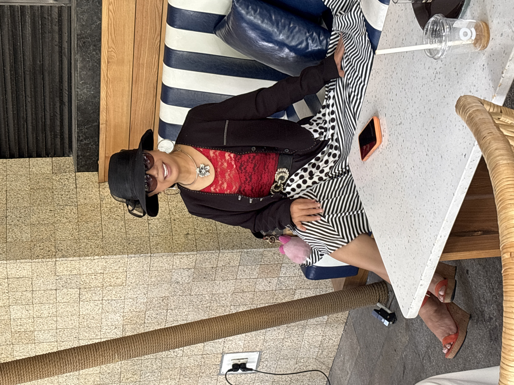 A Filipino woman in her 50s is seated on a bench in front of a building. She is wearing a black hat, sunglasses, a red lace top, a black jacket, and a black and white patterned skirt. She is also wearing orange platform sandals. The bench is blue and white striped. There is a table in front of her with a phone, a drink, and a straw. The building is made of light-colored brick. A rope-covered pole is to the left of the woman. The image was taken in front of the Vinpearl Luxury Nha Trang hotel in Nha Trang, Vietnam.