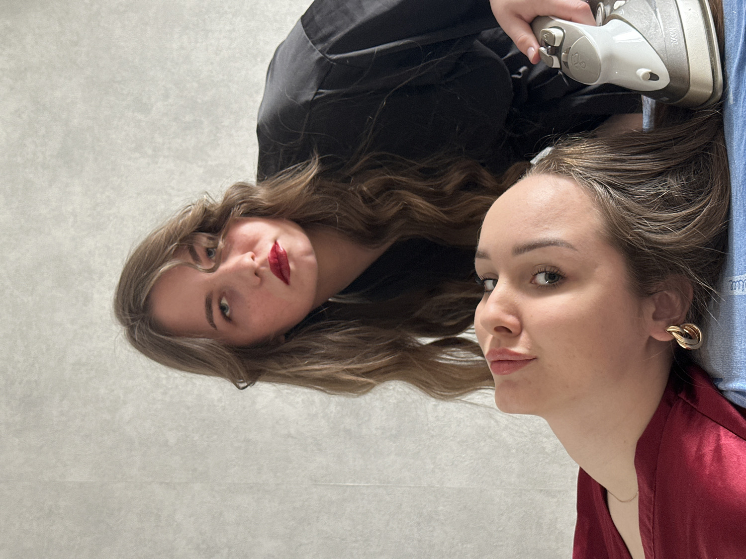 The image features two young women in an indoor setting, likely lying down against a light gray, textured surface resembling a wall or floor, in Chaikovsky, Russia. The photo's perspective is rotated, showing the women's heads oriented towards each other.
The woman on the left (when viewed upright) has long, wavy brown hair and wears a black top or dress. She looks directly at the viewer with a striking expression, enhanced by bright red lipstick.
The woman on the right has long, straight brown hair and a softer expression, also looking towards the viewer. She is dressed in a reddish, silky garment and sports a distinctive gold-colored, looped earring.
A silver and white clothing iron is prominently positioned above or near the right woman's hair, held by a hand. This appears to be a playful or artistic prop rather than actual use. The iron has "2000W" visibly printed on its side. A piece of blue denim fabric is also visible near the iron. The bright, even lighting suggests an indoor daytime shot.