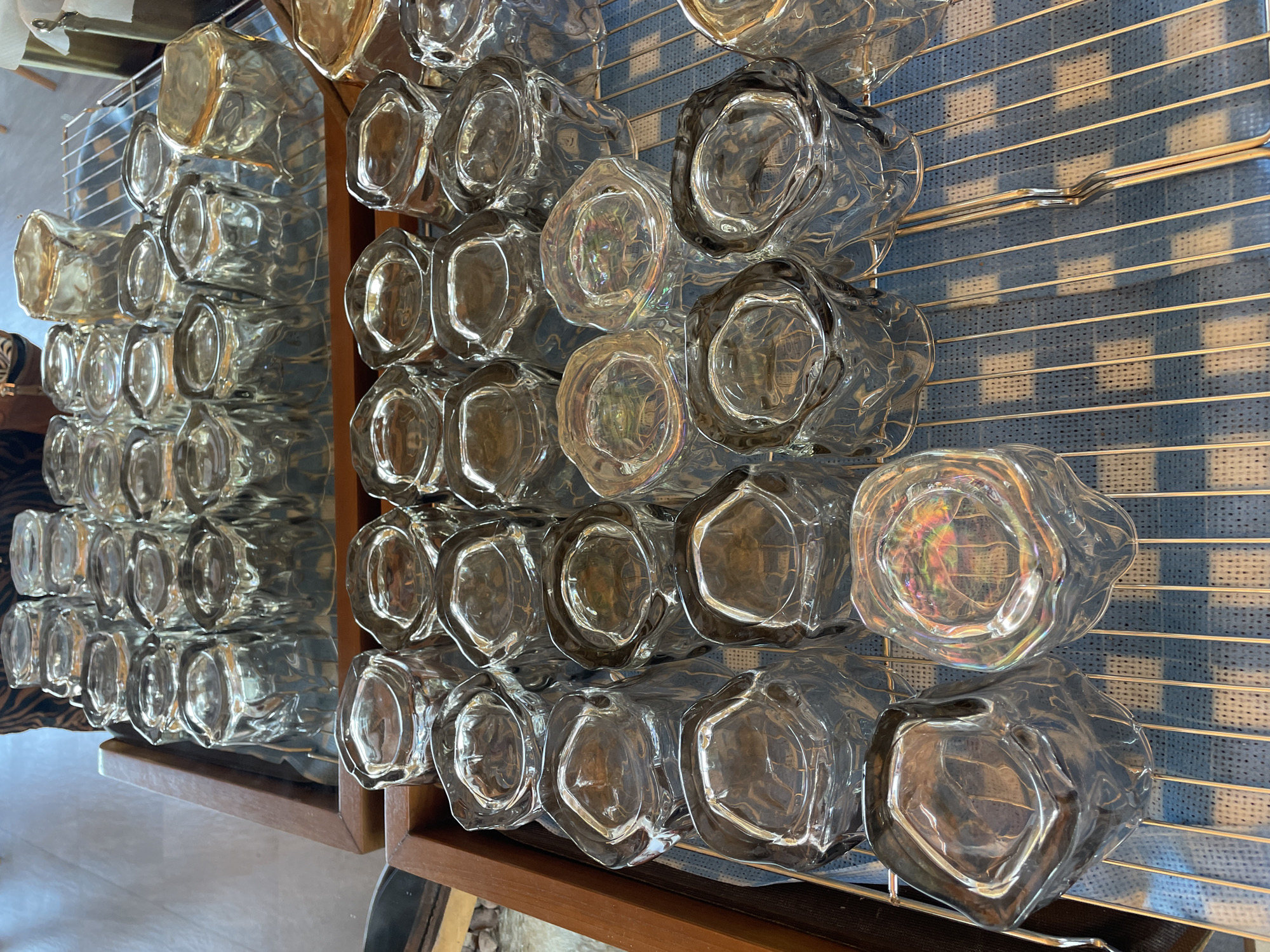 The image shows a collection of clear, textured drinking glasses displayed on shelves. The glasses have an unusual, somewhat organic shape, with slightly irregular edges. They appear to be made of thick glass, giving them a substantial and possibly weighty feel. The glasses are arranged in multiple rows, suggesting a storage or display area within a bar, restaurant, or retail establishment. The background is a blue and white checkered pattern. The image was taken indoors with bright lighting. The location is unknown.