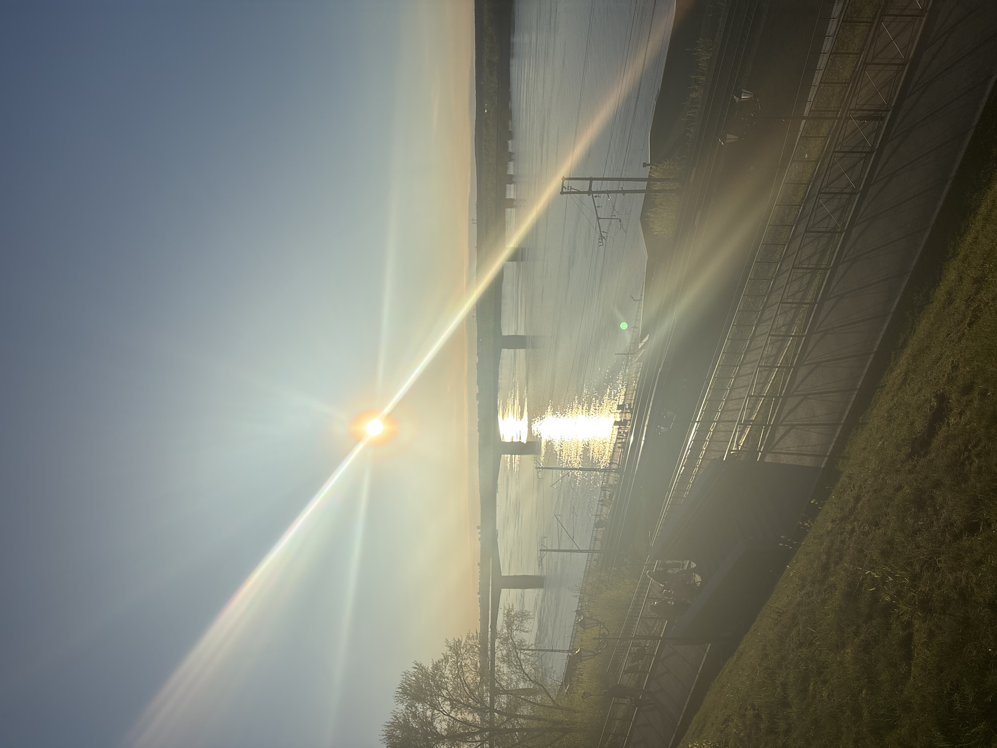 The image shows a horizontal view of a bridge over a wide river, likely in Perm, Russia, given the presence of a bridge and the river's width. The sun, the primary light source, is situated to the left of the bridge, its glare obscuring details of the bridge structure and the river's surface, which is reflecting light. Electrical support structures are visible along the shoreline, as well as a road, sidewalk, and grassy bank. The image is angled slightly to the right.