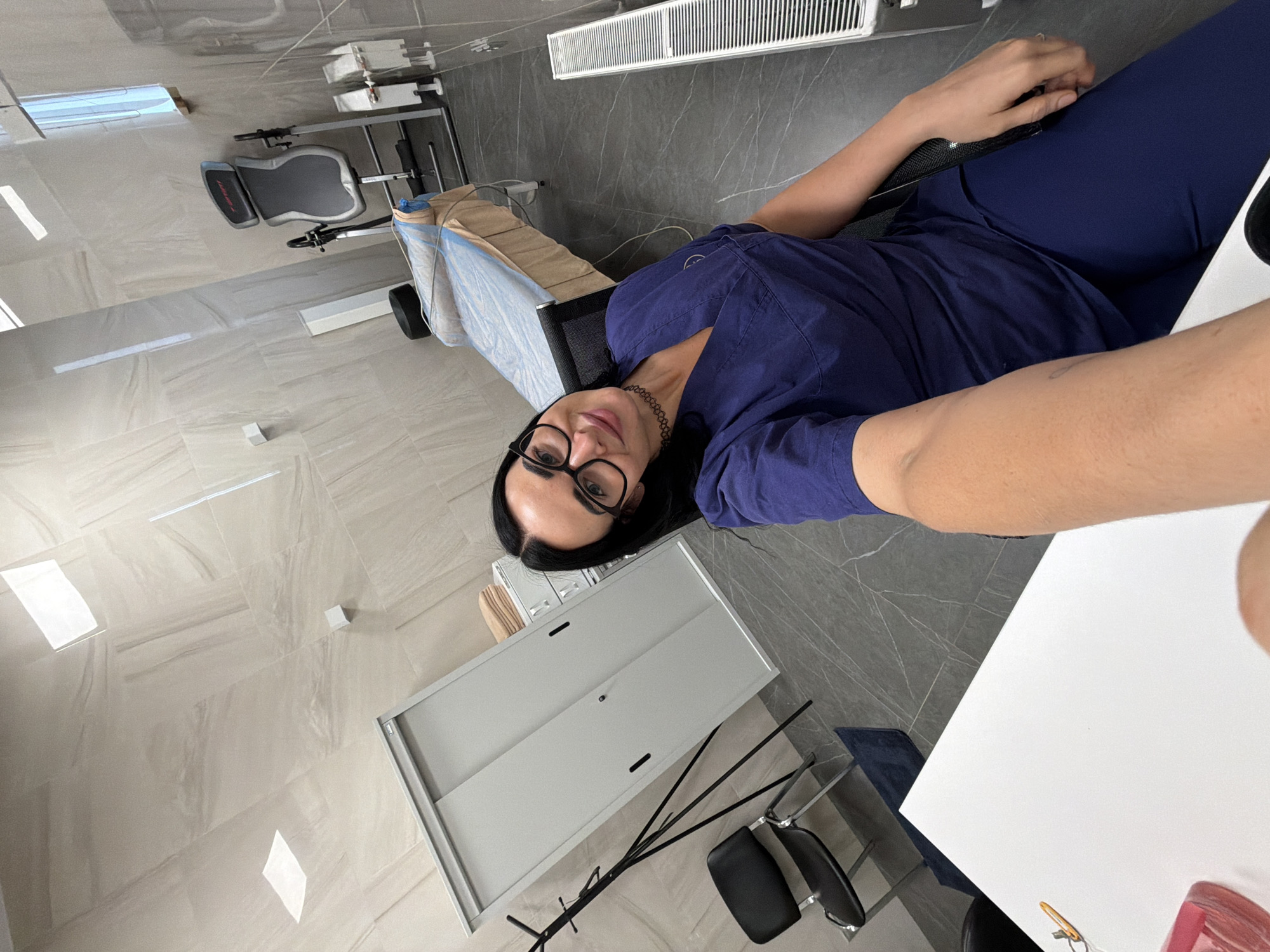A first-person perspective shot displays a person in blue scrubs reclining. The person wears glasses and a choker necklace. The location appears to be a medical facility, given the presence of medical equipment such as an examination table covered in a blue sheet. There's a white cabinet next to a modern black floor lamp. The floor is tiled in neutral shades of gray. The specific location in Saint Petersburg, Russia, cannot be determined from this image.