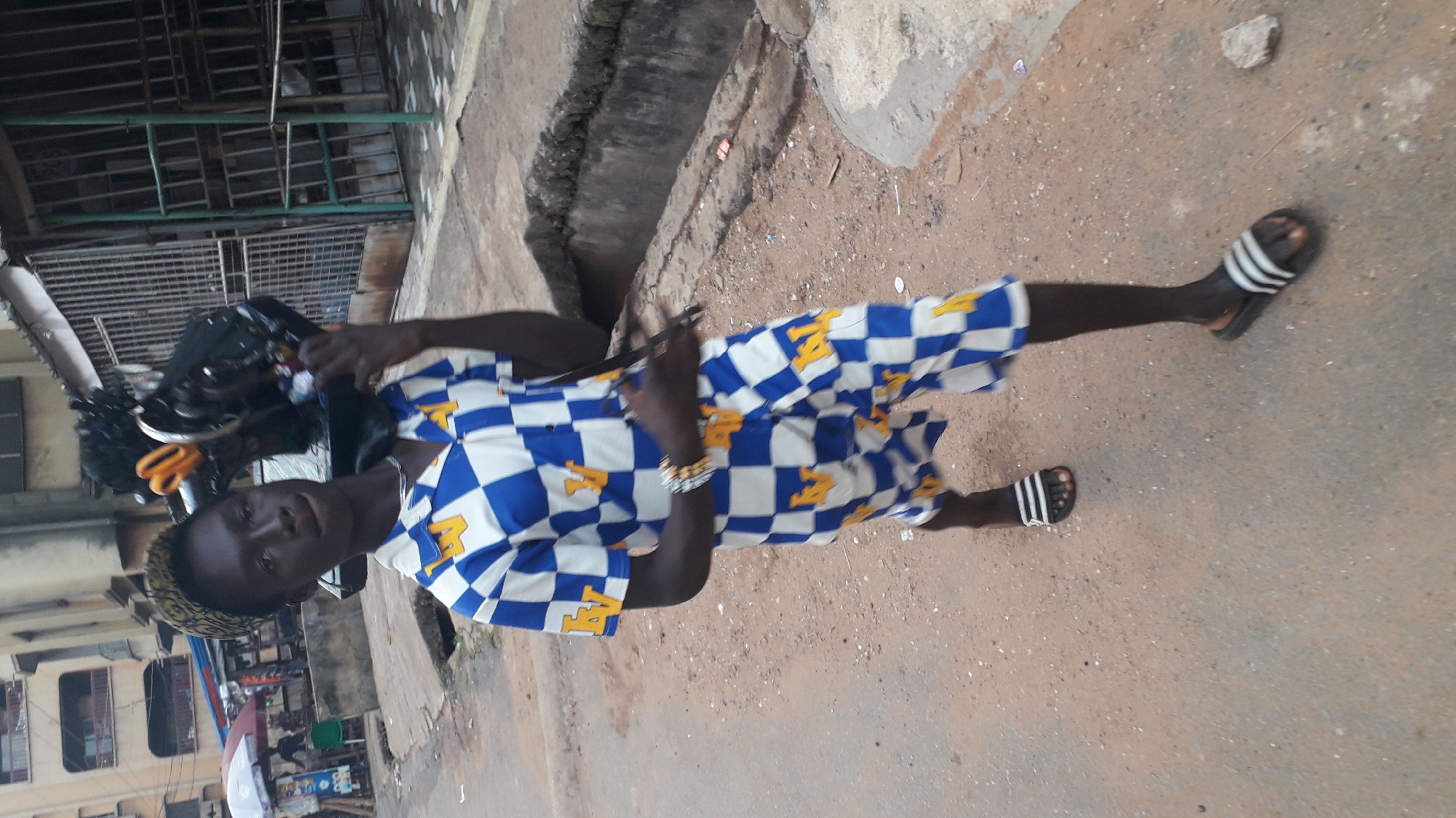 A person is standing in a street in Onitsha, Nigeria. The person is wearing a blue and white checkered shirt with a yellow design, black Adidas slides, and is holding a black object over their shoulder. The street is paved, and there is a building in the background. Other items of note include buildings, a sewage ditch, an umbrella, a market, and metal security bars.