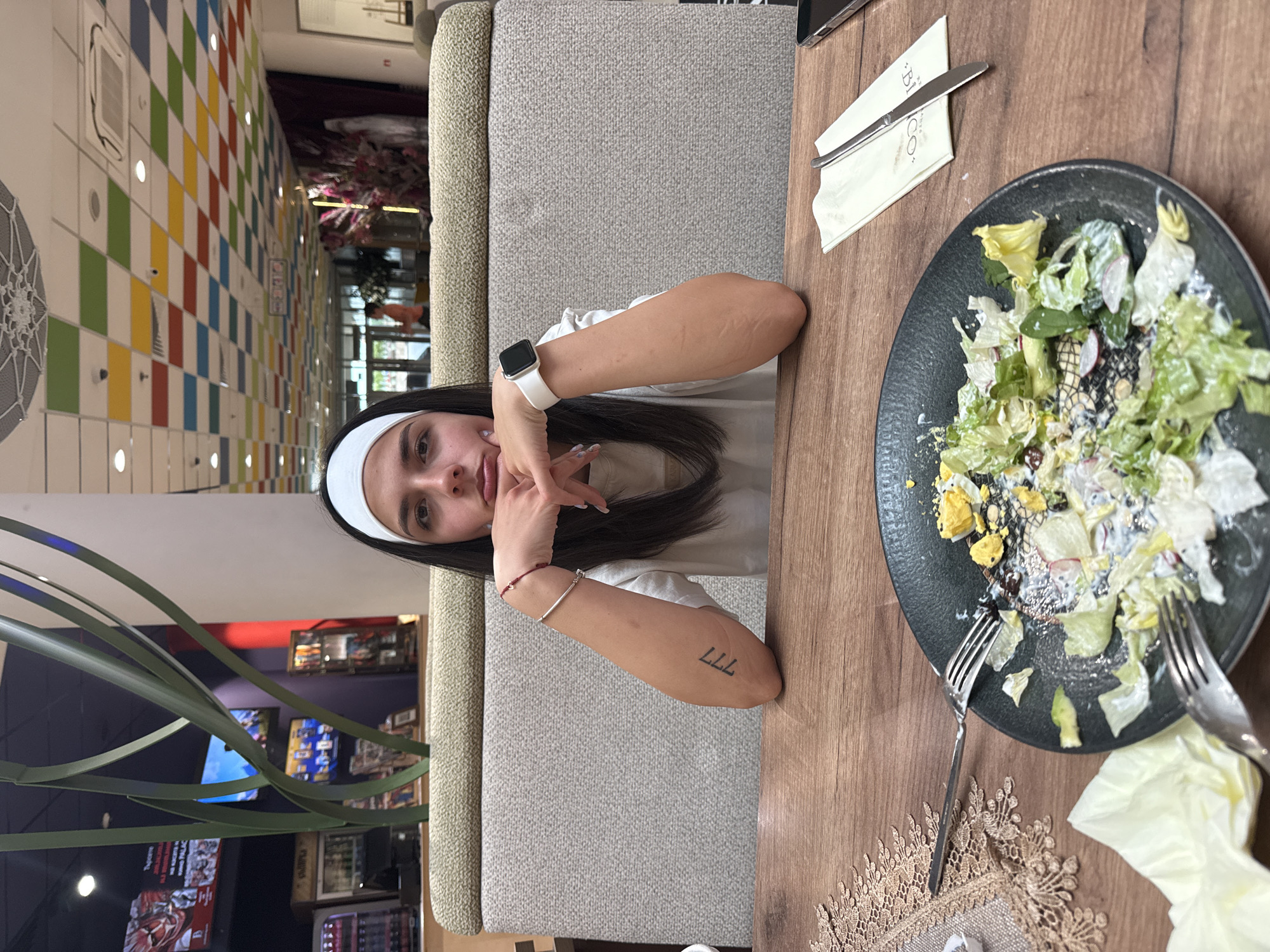 The image depicts a young woman seated at a wooden table in what is identified as a restaurant or food court in Veliko Tarnovo, Bulgaria. She is positioned centrally, looking directly at the camera with a contemplative expression, her chin resting on her clasped hands. She has long dark hair and wears a white headband and a light-colored short-sleeved top. On her left wrist, she wears a silver bracelet and a red string bracelet, while her inner left forearm bears a "777" tattoo. On her right wrist, she wears a white smartwatch with a dark screen; the skin on her right forearm exhibits multiple faint, linear scars.
In front of her on the wooden table, a dark, round plate holds the remnants of a largely consumed salad, featuring lettuce, white dressing, radish slices, and pieces of hard-boiled egg. Two silver forks are near the plate, one partially resting on it. To the left of the plate, a crumpled light-yellow paper napkin is visible, alongside a decorative, lace-patterned beige table runner. To the right, a silver knife rests on a folded white paper napkin with "BICO." and "BAR" printed on it. A dark smartphone is partially visible on the far right edge of the table.
The woman is seated in a booth-style bench with a light grey textured fabric backrest and an olive-green top cushion. The background shows a modern indoor environment. Behind her, a large, abstract artificial plant with green, blade-like leaves is visible. Further back, two flat-screen televisions display what appears to be content with Cyrillic text. The ceiling is distinctive, featuring a checkerboard pattern of square panels in various colors (red, yellow, green, blue, white), interspersed with recessed lighting and air vents. A circular, white woven ceiling medallion is also visible. Security cameras are mounted on the ceiling. In the distant background, a brightly lit area with additional seating and decorative elements, possibly including artificial flowers, can be seen, consistent with a commercial establishment.
