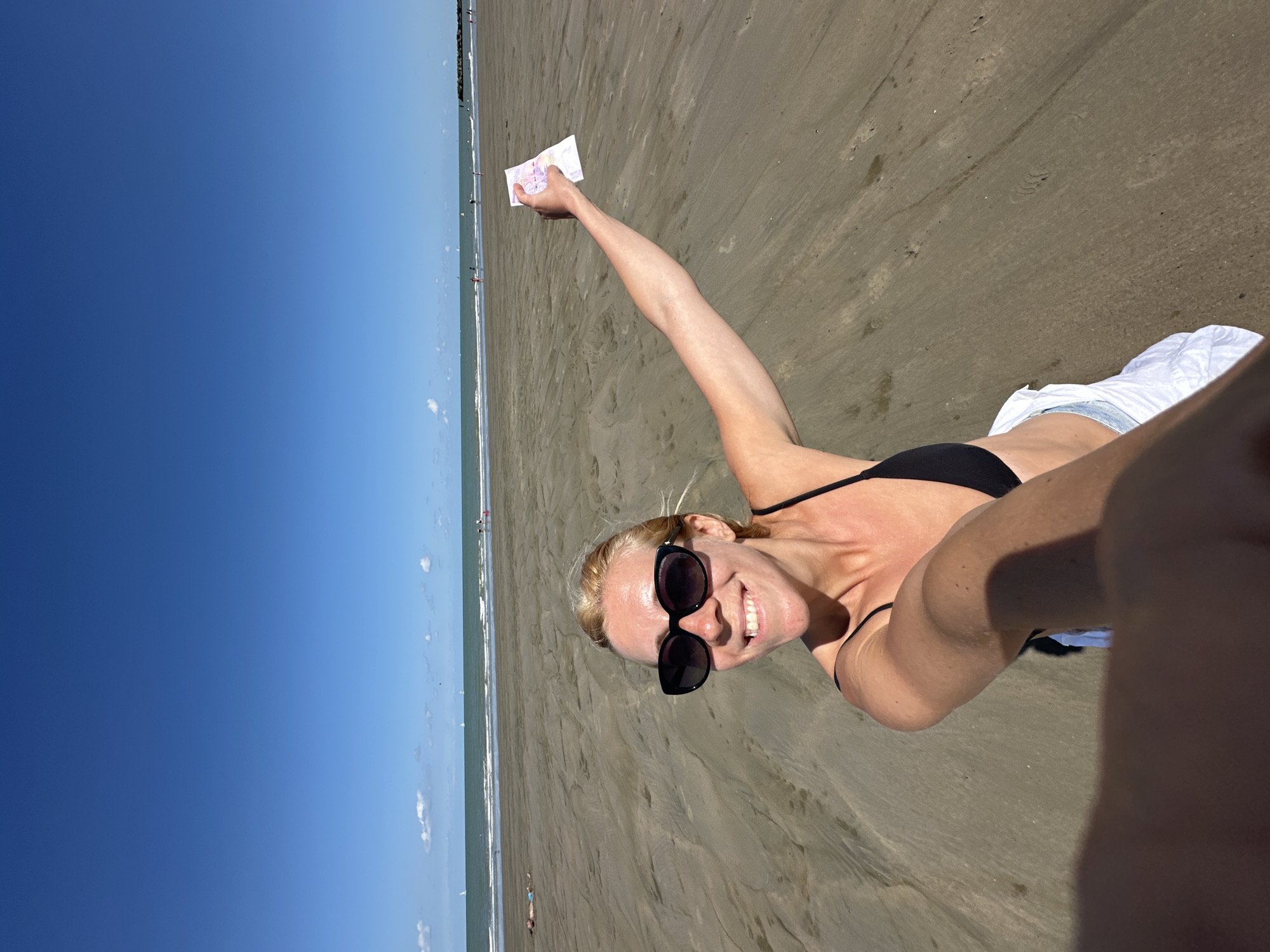 A woman with blonde hair, wearing a black bikini top and sunglasses, smiles at the camera while holding a white piece of paper. She is on a sandy beach, with the ocean and a clear blue sky in the background. Several sailboats are visible on the water, and a few people are scattered along the shoreline. The scene is indicative of a sunny day at the beach, likely in a coastal area. Dunkirk, France, is a coastal city known for its beaches, which could be the location depicted.