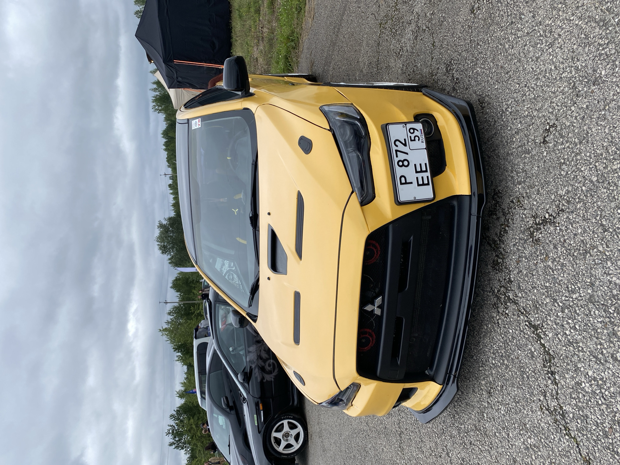 An eye-level, full frontal medium shot captures a yellow Mitsubishi Lancer Evolution X parked on an asphalt surface. The car's black grille features two red accents and the Mitsubishi emblem. A black lip spoiler is attached to the front bumper. The license plate, P 872 EE 59 RUS, is clearly visible. The car has a black roof and a black side mirror. In the background, other cars, including a black Mitsubishi Lancer, are parked. Several black tents and some trees are also visible. The sky is overcast with gray clouds. The scene appears to be at an outdoor event or car meet in Perm, Russia.