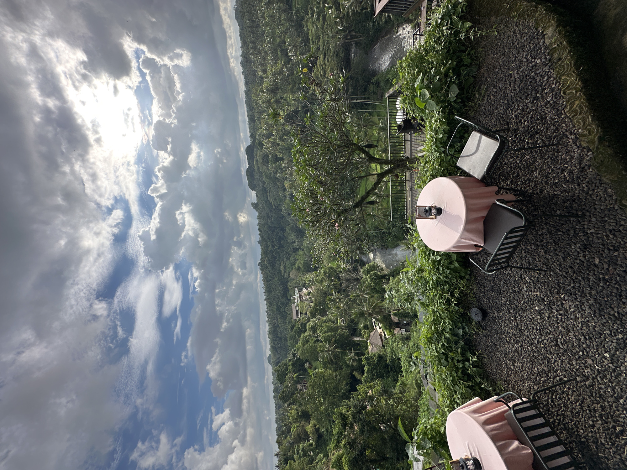 The image shows an outdoor cafe setting in Ubud District, Indonesia. There are tables with pink tablecloths and metal chairs arranged on a gravel surface. The cafe overlooks a lush, green valley filled with trees and foliage. In the background, a cloudy sky dominates the scene.
