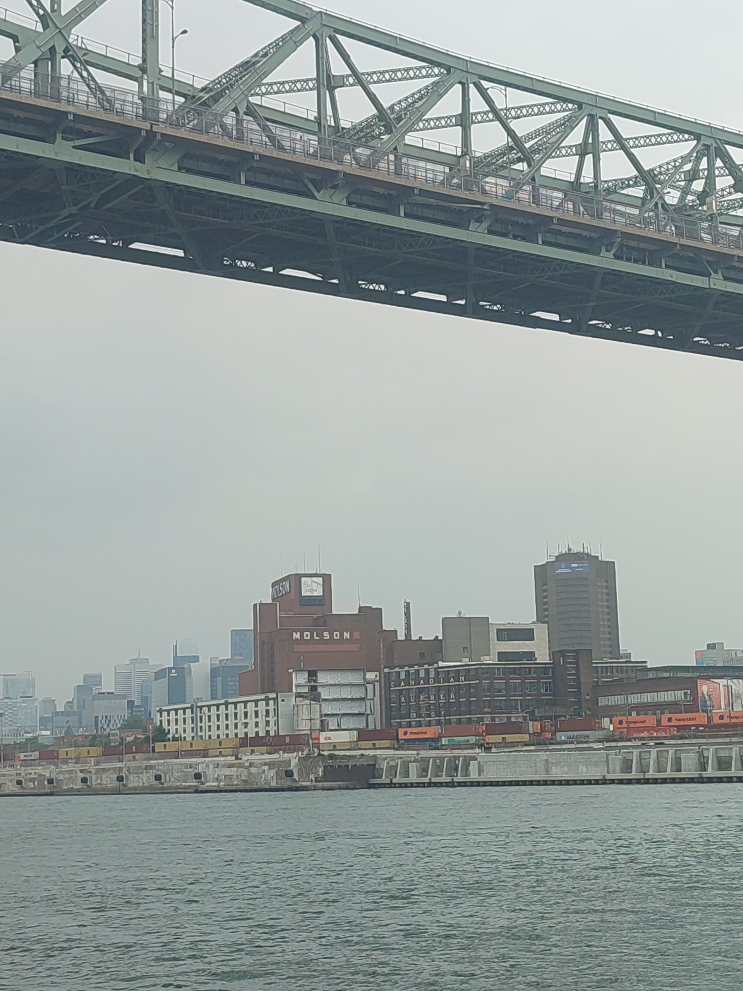 The image captures a view of the Montreal waterfront, likely from the St. Lawrence River, under a hazy sky. Dominating the upper portion of the frame is the underside of a large, green metal truss bridge. Below the bridge, the city skyline is visible, featuring a mix of industrial buildings and modern high-rises.
Prominently featured is a large, red brick building with a prominent "MOLSON" sign, indicating the presence of the historic Molson Brewery or related facilities. Adjacent to it are other industrial structures, including warehouses and what appear to be shipping containers stacked along the waterfront. A tall, slender skyscraper stands further in the background, characteristic of Montreal's downtown core.
The water in the foreground is a murky green-blue, with gentle ripples suggesting calm conditions. The overall atmosphere is overcast and slightly muted, characteristic of a foggy or hazy day. No people are clearly discernible in the image, though figures might be present on the bridge or along the waterfront, too distant or obscured to identify. The scene depicts a blend of industrial heritage and urban development along the waterfront of Montreal.