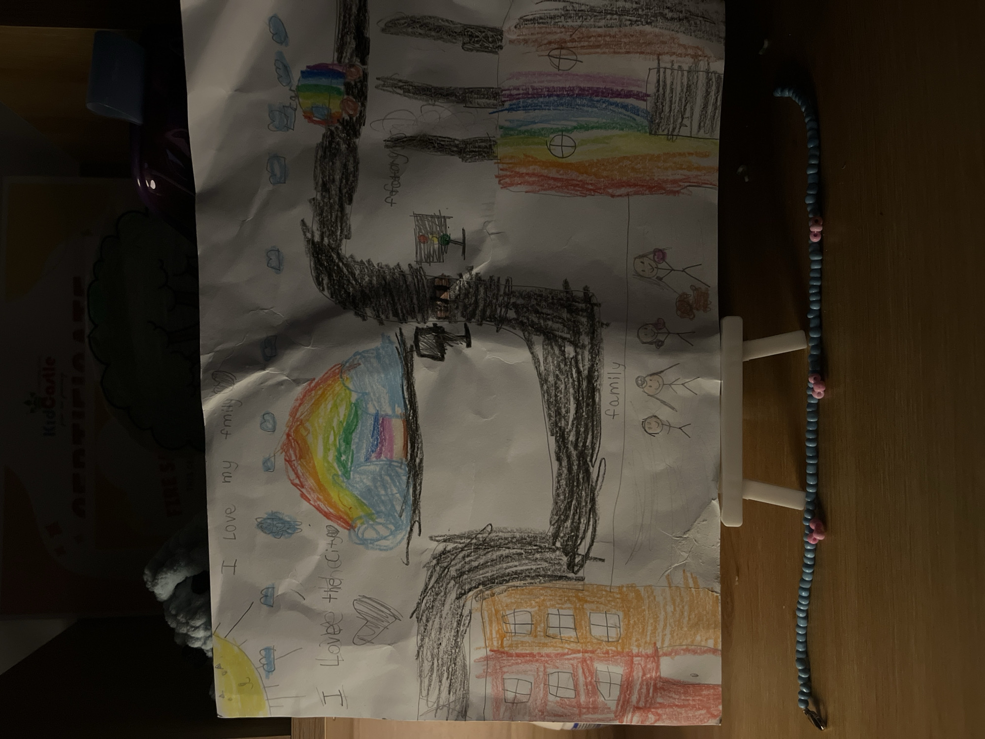 A child's drawing on a white sheet of paper depicts a cityscape. The drawing includes representations of buildings, a rainbow, clouds, the sun, and stick figures representing people. Text within the drawing includes phrases like "I love my family" and "I love the city." The drawing is positioned on a small, white easel, placed on a wooden surface. A beaded necklace is also on the surface next to the drawing. The surrounding environment includes a shelf with various items, including a book or certificate with the words "KidCastle" visible. The location in the city of Bac Nha Trang Ward, Vietnam, is not directly evident from the image.