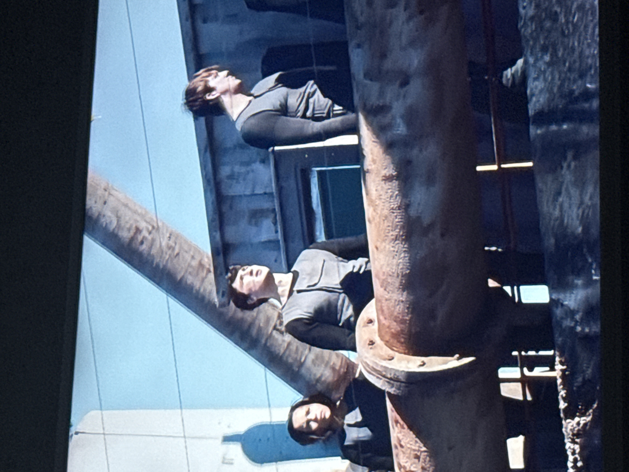 The image depicts three people in what appears to be an industrial or urban setting. The perspective is unusual, seemingly angled upwards. The figures are dressed in similar, utilitarian clothing, suggesting a uniform or work attire. One person is looking directly at the camera, while the other two appear to be looking upwards or towards the sky. They are positioned around or near large, rusty pipes and metal structures. The background shows glimpses of blue sky and white or light-colored building facades, indicating an outdoor location. The city of Mytishchi, Russia, is not discernible from the image alone. Further information or context would be needed to confirm a specific location.