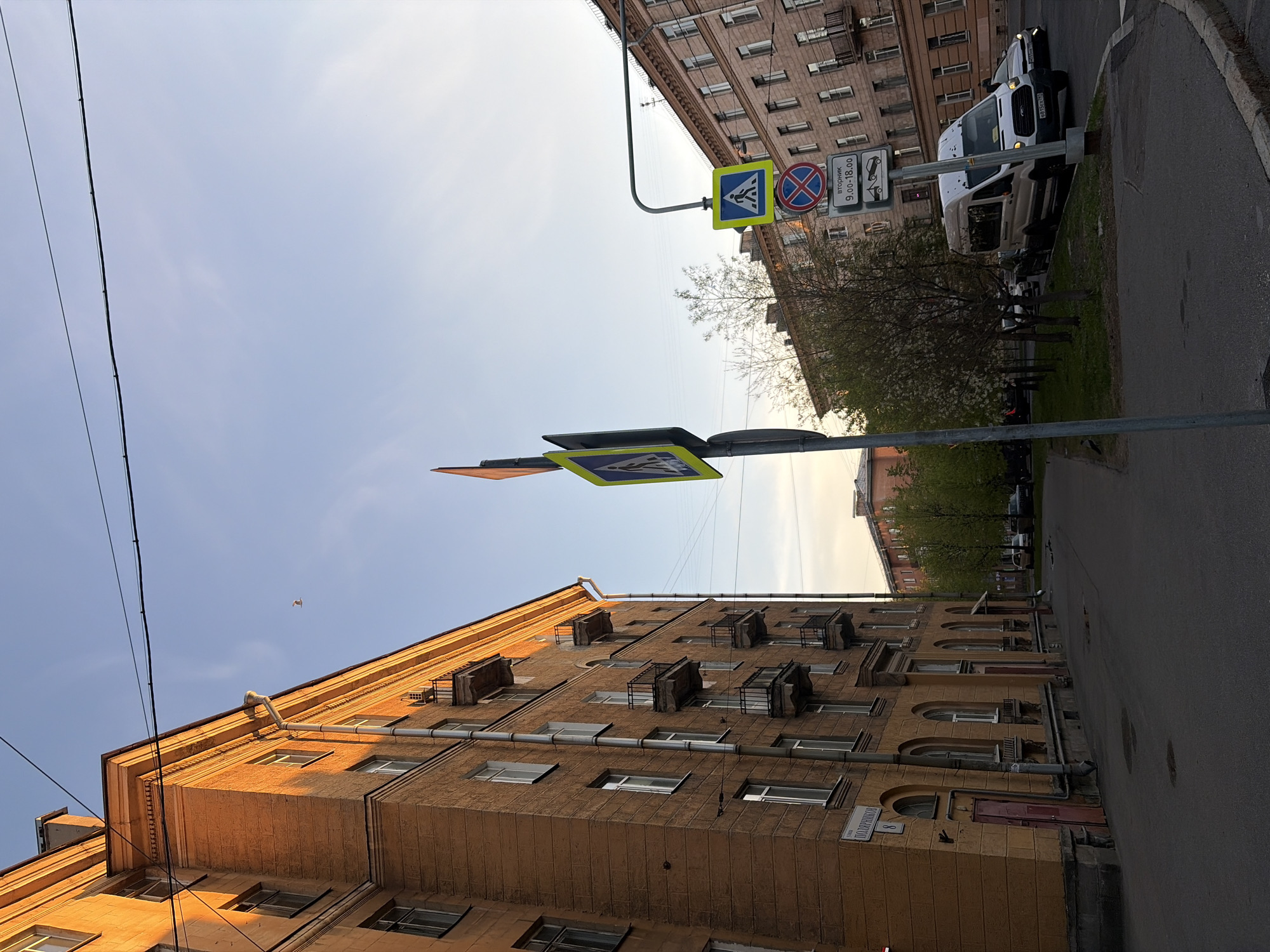 The image depicts a street scene in Saint Petersburg, Russia, looking up towards the sky. The shot is taken at street level, with buildings on either side. The building on the left is a multi-story structure with light-brown facade, with rows of windows and protruding balconies. A street sign with the name "ПОЛОЗОВСКАЯ" (Polozovskaya) is visible on the lower right.
To the right, another building can be seen, lighter in color, with a row of parked vehicles.
In the center of the image, there are two pedestrian crossing signs on poles. The sky is visible above the buildings, with some power lines. The light suggests it's daytime.
