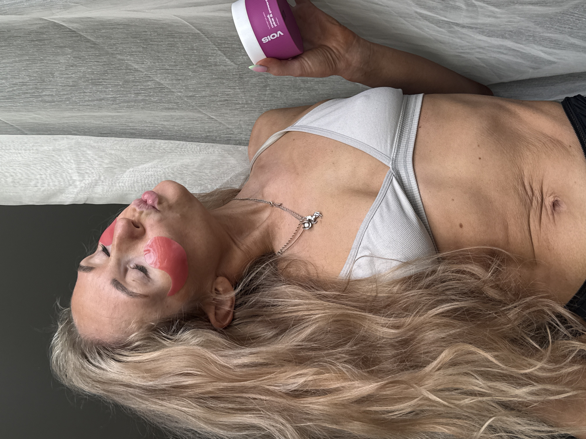 This is a vertically oriented, medium-shot photograph of a woman lying down with her head tilted back. She has long, wavy blonde hair that cascades around her face and shoulders. She is wearing a light gray sports bra and black underwear. On her cheeks, under her eyes, are two pink under-eye patches. She is holding a round, purple jar with a white lid in her right hand. The text on the jar reads "VOIS" in white capital letters. Below that, in smaller white text, it says "mise: pink & power" and "collagen".
The woman appears relaxed, with her eyes closed and her mouth slightly open. The lighting suggests it is daytime, possibly indoors, given the softness of the light. The background consists of two textured surfaces: a lighter, rippled fabric above and a darker, solid surface below. The overall impression is one of self-care or relaxation. The context of Chaikovsky, Russia is provided but not visually evident in the image itself.