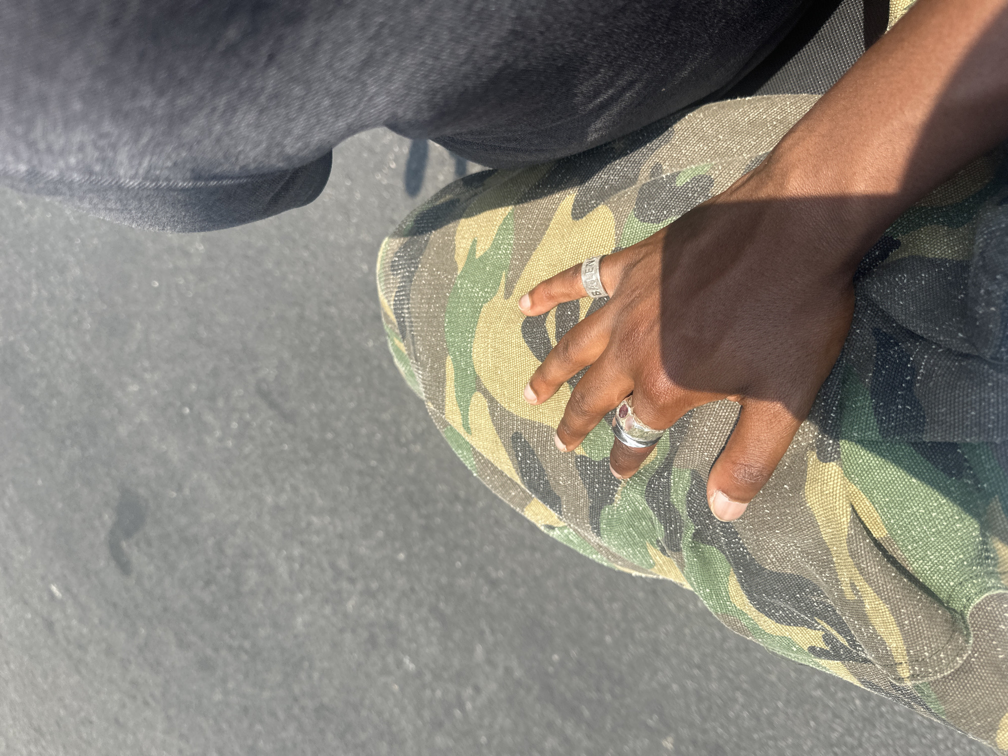 An overhead shot, taken in Bangkok, Thailand, captures a person's right hand resting on a camouflage-patterned item or garment. The individual, whose skin tone is dark, is partially visible, with their hand and a portion of their legs or lap in view. The upper left of the frame shows dark, possibly black, fabric, likely part of the person's pants or jeans.
The hand wears two prominent silver-colored rings. On the ring finger, a band features the clearly legible text "BALEN," suggesting the brand Balenciaga. Another ring on the pinky finger appears to have a small, red or purple gemstone.
The scene is brightly lit by strong sunlight, which casts a distinct, sharp shadow of the hand across the camouflage pattern, indicating a clear, sunny day, likely around mid-day. The ground surface, visible in the lower left, is grey and textured, resembling asphalt or concrete. No specific activity or emotion is discernible beyond the hand's resting position.