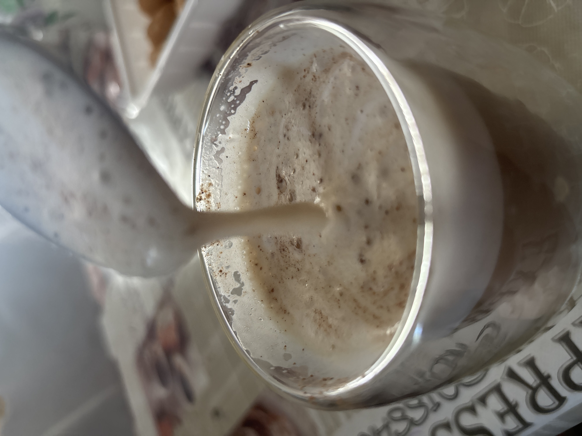 A close-up, low-angle shot shows a clear glass mug with a double wall, filled with a frothy, light brown beverage, likely coffee or hot chocolate, being poured from above. The beverage has a foamy top with small brown flecks, possibly cinnamon or coffee grounds. The mug rests on a patterned tablecloth. In the background, a white plastic container with some golden-brown food items, possibly pastries or cookies, is visible. The image is taken indoors, possibly in a home or cafe setting, with soft, natural lighting. The scene does not provide any specific information or context about the city of Zubov, Russia.