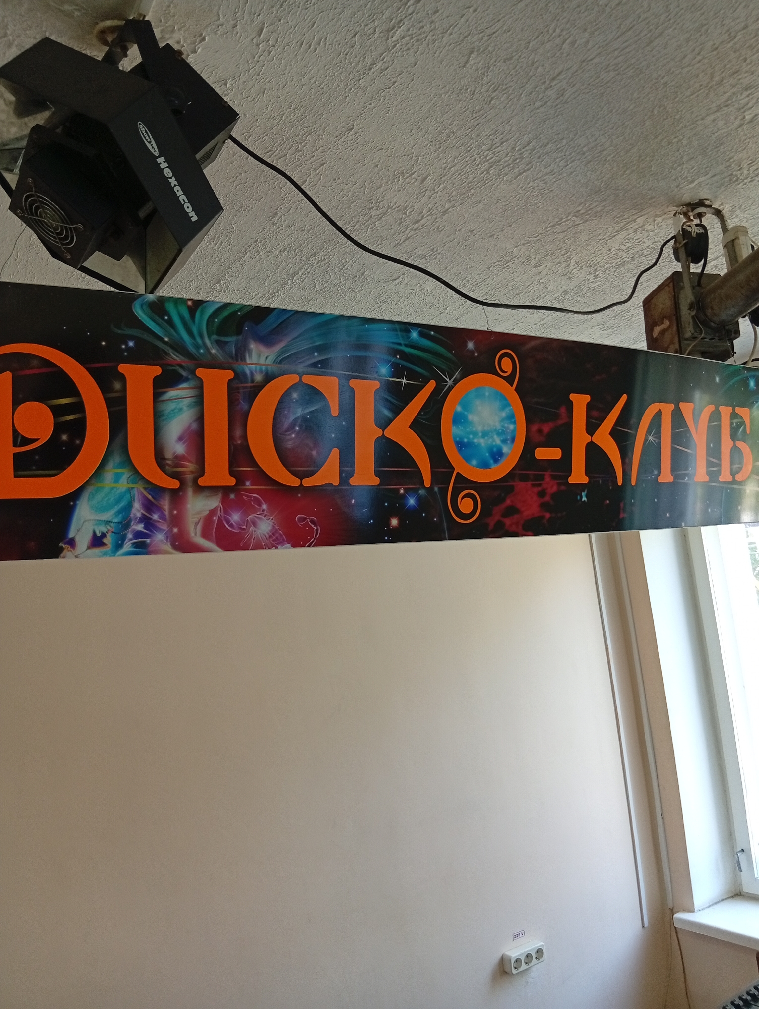 A close-up, slightly angled shot shows an orange sign with the Cyrillic text "ДИСКО-КЛУБ" (Disco Club) against a dark background with abstract, colorful imagery of what appears to be nebulae, stars, and a scorpion. The sign is mounted on a wall or ceiling. Above the sign, on the textured ceiling, are two black stage lighting fixtures. One fixture, labeled "Showtec Hexacon," is visible on the left, featuring a fan. To the right, a section of a metal pipe and some electrical components are mounted. Below the sign, the wall is a plain, light beige color. In the lower right corner, a white electrical outlet with three sockets is visible on the beige wall, labeled "220 V". The overall scene appears to be indoors, possibly in a club or entertainment venue, in Крыўлянскі сельскі Савет, Belarus.