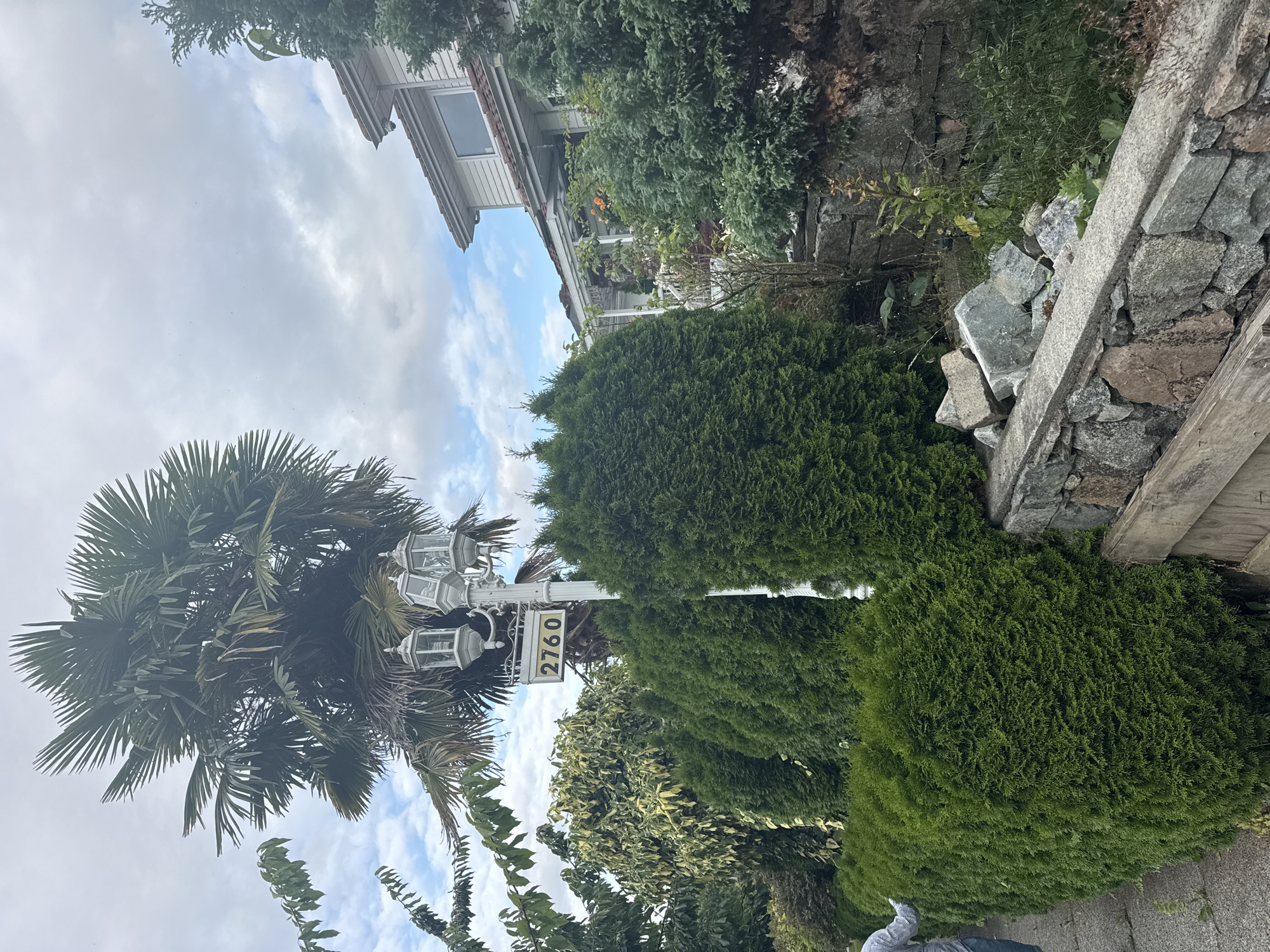 The image shows a residential house with a palm tree and manicured hedges. A street lamp with three lights is visible, along with a sign displaying the number "2760". A person wearing a blue and white checkered shirt and blue jeans is standing on the left side of the frame, near a sidewalk and a stone wall. The house appears to be a two-story structure with a white exterior and a brown roof. The sky is cloudy, with patches of blue sky visible. The overall impression is of a suburban street in a temperate climate.