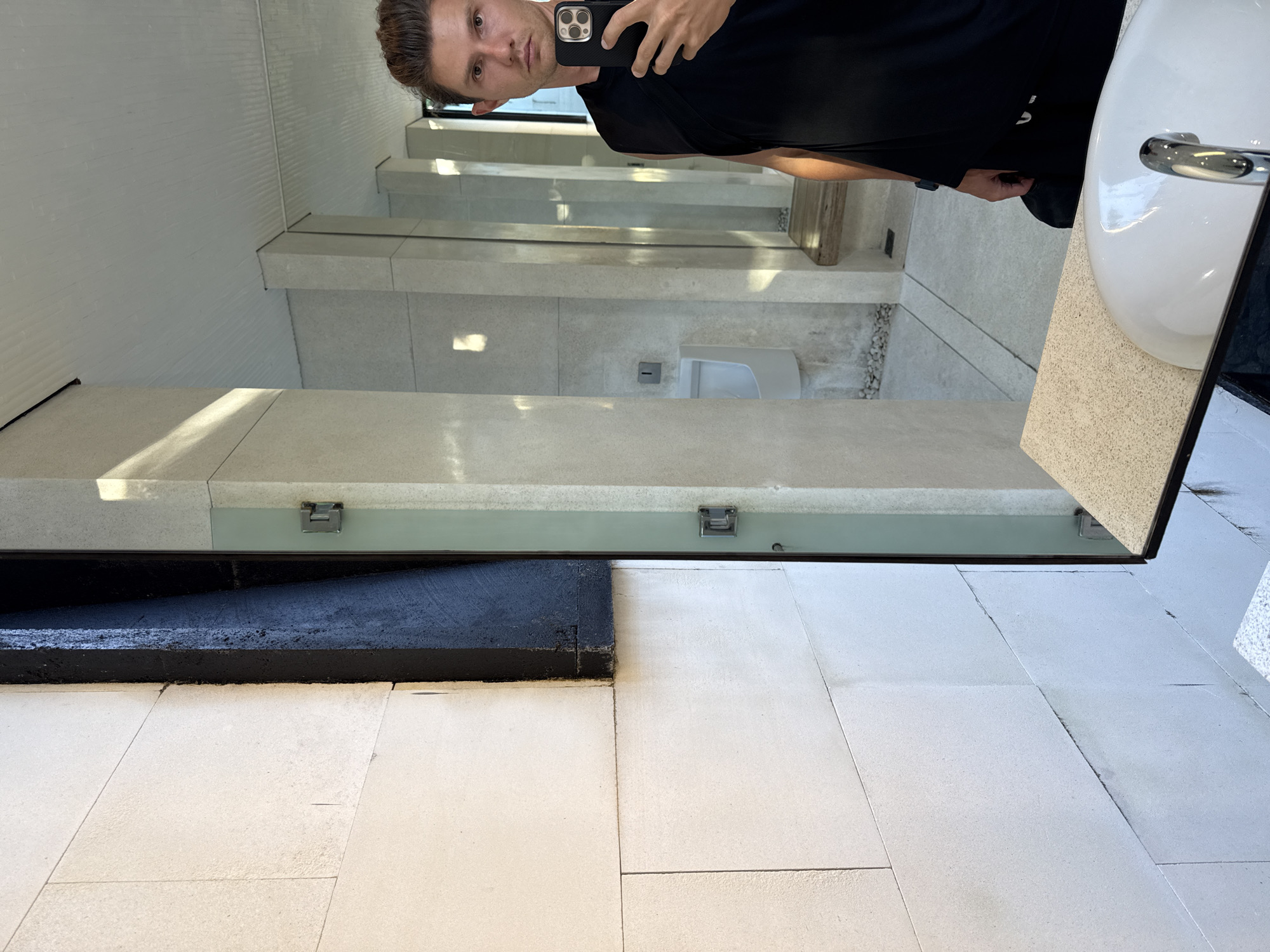 The image is a mirror selfie taken in a bathroom in Kuta Selatan, Indonesia. The individual taking the photo is a light-skinned male with short brown hair, wearing a black t-shirt. He is holding a black smartphone with a dual-lens camera in his right hand.
The bathroom features a large mirror reflecting the room's interior. Reflected details include a white, wall-mounted urinal, concrete-like countertops, and white tiled floors. Below the mirror is a dark gray or black ledge. A white porcelain sink with chrome fixtures is visible on the right side of the mirror, close to the photographer. The overall impression is a modern, minimalist bathroom design.