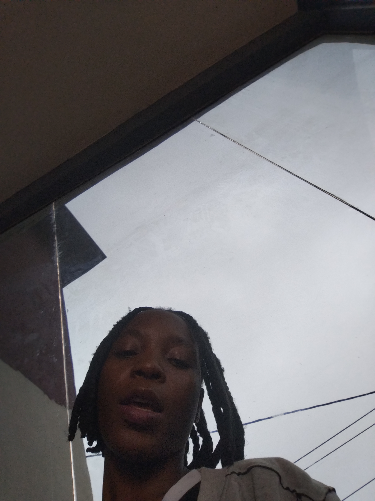 A low-angle, medium shot captures a young Black woman with dreadlocks standing outdoors against a grey, overcast sky. She is looking up and slightly to the left, her mouth slightly open. She is wearing a grey shirt with a dark, wavy trim along the neckline. The background is dominated by a large window or glass panel reflecting a building structure to the left and power lines against the sky. The scene appears to be taken in Uselu, Nigeria, likely during the daytime.