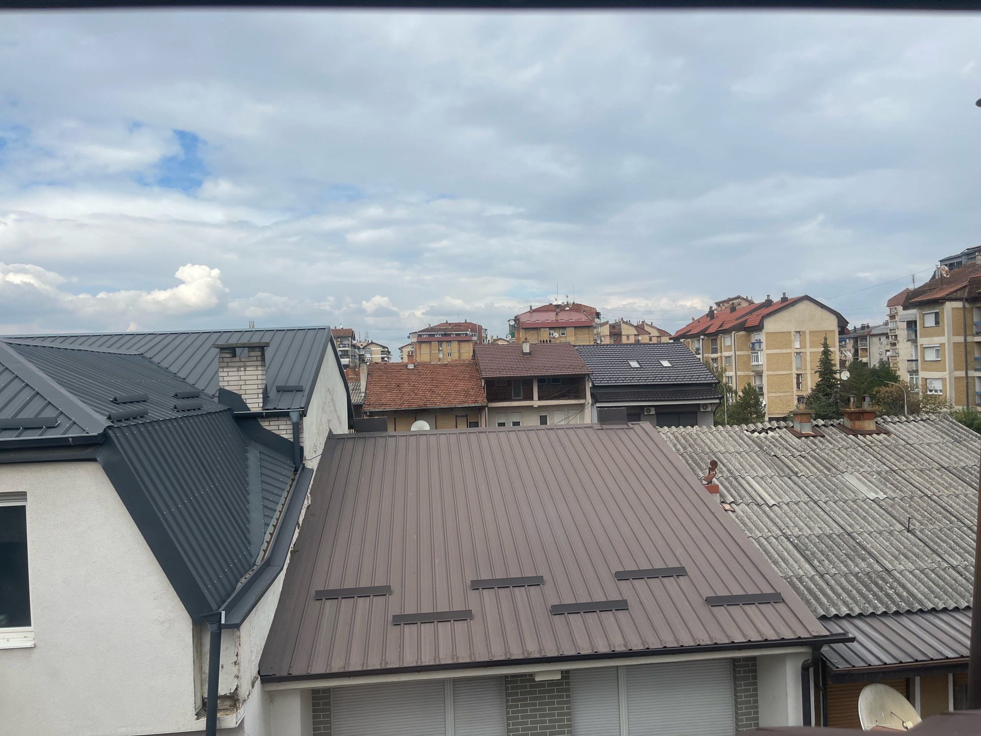 The image shows a view of rooftops in Prizren, Kosovo, under a cloudy sky. The foreground features several houses with various roofing materials. On the left, a white house has a dark gray corrugated metal roof. To its right, a house with a dark brown metal roof is partially visible. In the center, a large house has a brown metal roof with several black horizontal slats. To the right of this, a house is covered with corrugated asbestos tiles, showing signs of weathering and some discoloration. In the background, a cityscape with residential buildings and apartment blocks is visible, with some trees interspersed. The sky is filled with a mix of white and gray clouds, with patches of blue sky showing through. There are no people or animals visible in the image. The scene appears to be a typical urban residential area.