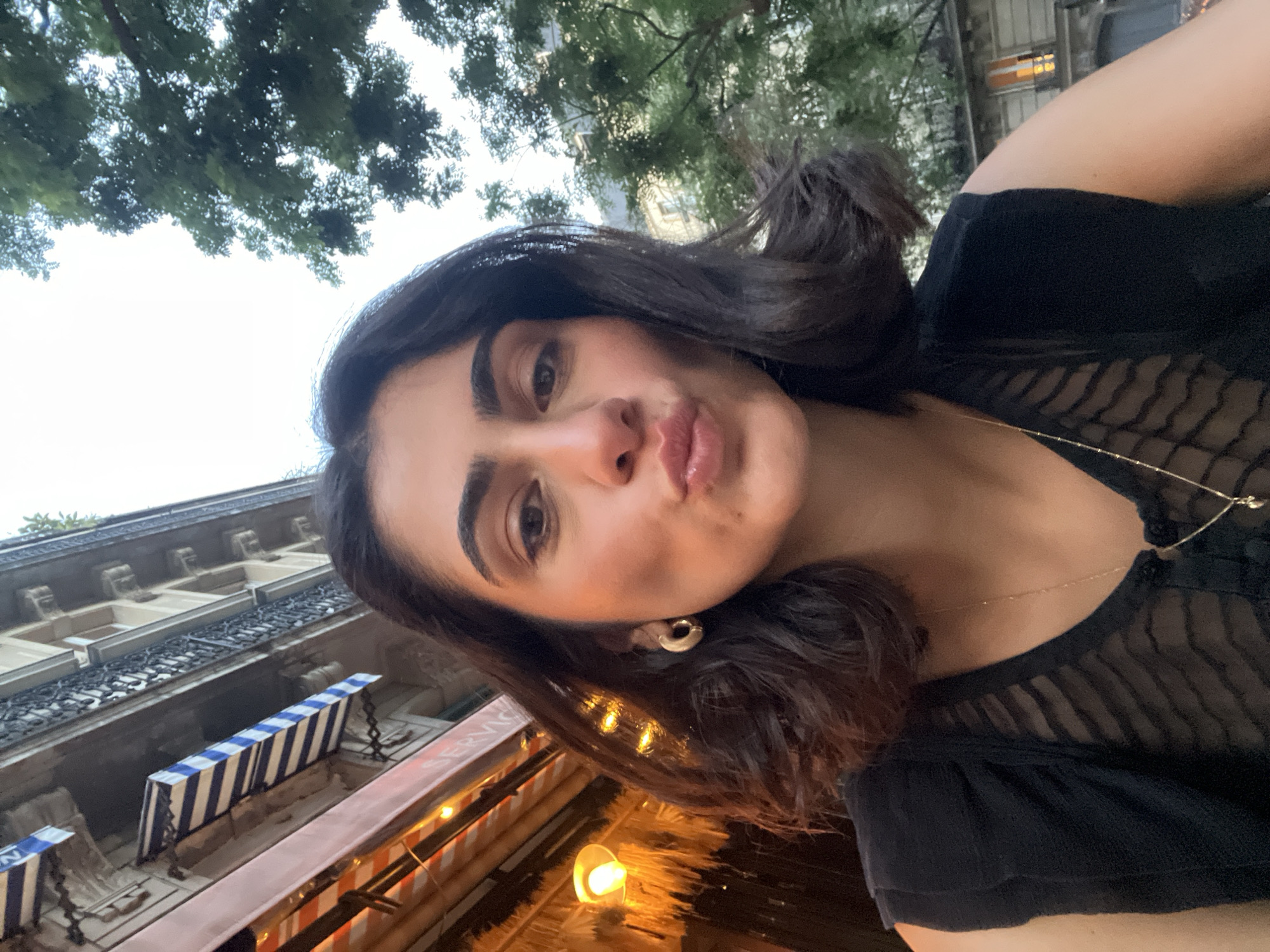 A young woman with dark, wavy hair is taking a selfie outdoors in Paris, France. She is wearing a black sleeveless top with a sheer, patterned fabric over the chest and a delicate gold necklace. A gold hoop earring is visible on her left ear. Her lips are puckered, and she is looking directly at the camera. In the background, a building with awnings and balconies is visible, along with lush green trees. The lighting suggests it is daytime, possibly late afternoon or early evening.
