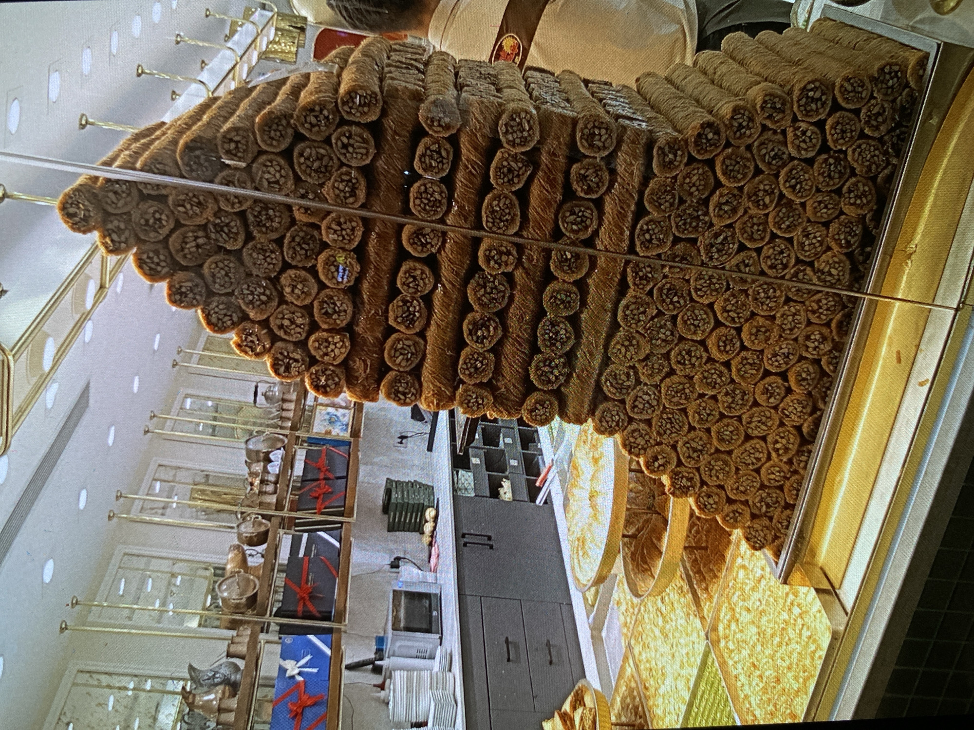 This vertically-oriented image captures a vibrant display of traditional sweets within what appears to be a confectionery shop or patisserie in Bankya, Bulgaria.
The central and most prominent feature is a massive, meticulously stacked arrangement of cylindrical pastries. These individual rolls, rich brown in color, are filled with finely chopped nuts (likely pistachios or walnuts), visible at their cut ends, suggesting a type of baklava such as *sarma* or *finger baklava*. They are stacked in an impressive, almost pyramid-like formation behind a glass barrier, inviting customers to view them from various angles.
Below and to the right of this large display, several trays hold other varieties of golden-hued sweets, some appearing flat and rectangular, others round, with crumbly or shredded textures, possibly other forms of baklava or kadaif.
In the background, on the left side of the image, there are elegant display cases with glass shelves. These shelves showcase decorative items including small brass-colored pots and elegantly wrapped gift boxes. Some boxes are dark blue or black with red ribbons, while one is light blue featuring a white bird-like design and a red ribbon. The walls are light-colored with multiple recessed oval lights.
On the far right in the background, a person's upper body is partially visible, wearing a light-colored uniform or apron with a small, indistinct emblem on the shoulder. Further back on the right, there's a functional counter area with a dark grey cabinet, a microwave, and other storage items.
The overall scene depicts a well-stocked and inviting shop specializing in delectable pastries, likely catering to customers looking for traditional Bulgarian or Balkan sweets. No clear, legible text is visible in the image.