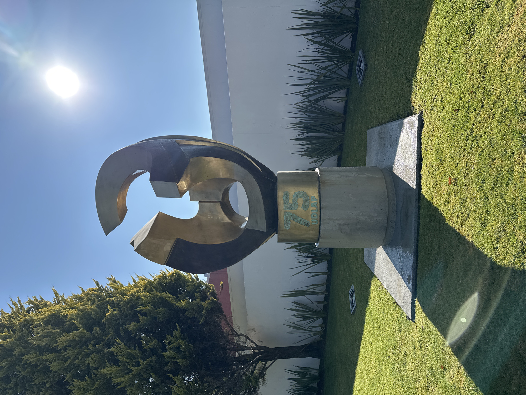 The image depicts an outdoor scene in San Andrés Cholula, Mexico, centered on a large, abstract sculpture. The sculpture, crafted from golden-bronze metal, features an irregular, multifaceted design composed of both curved and angular segments. It stands atop a cylindrical concrete pedestal. A golden band encircles the upper part of the pedestal, displaying the green inscription "75 UDLAP," identifying it as being on the campus of the Universidad de las Américas Puebla. The sculpture is situated on a well-maintained green lawn. Behind it, a row of spiky green plants lines the base of a long, white wall. A tall, dark green coniferous tree stands to the left of the sculpture. The sky above is clear and bright blue, with the sun appearing as a brilliant orb in the upper right quadrant, casting distinct shadows across the lawn and the sculpture's concrete base. A square metal grate is visible, embedded in the grass in the lower mid-ground. No individuals are present, and no actions or interactions are depicted.