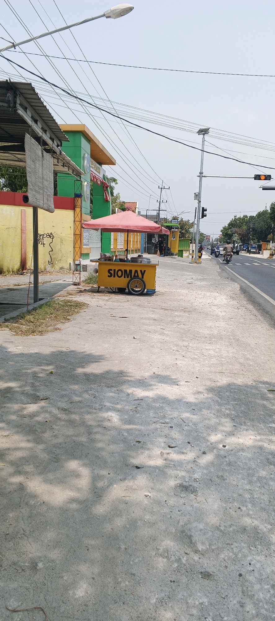 This image captures a street scene in Glogok, Indonesia. In the foreground, a sidewalk made of rough concrete is visible, with scattered leaves and debris. The sidewalk is partially shaded by dappled sunlight filtering through trees. To the right, a paved road runs parallel to the sidewalk. On the left side of the image, a building with a yellow and green facade stands. A portion of a covered structure with a corrugated metal roof is also present.
Dominating the center of the image is a bright yellow food cart with a red umbrella. The word "SIOMAY" is prominently displayed in black letters on the cart. The cart has wheels and appears to be stocked with food items. Behind the food cart, there are some people and other structures, including a yellow booth or small building.
Further down the road, traffic signals are visible, with one displaying an orange light. Cars and motorcycles are present on the road, indicating ongoing activity. The sky is clear and bright. The overall impression is that of a typical roadside scene in an Indonesian city, likely during the daytime.