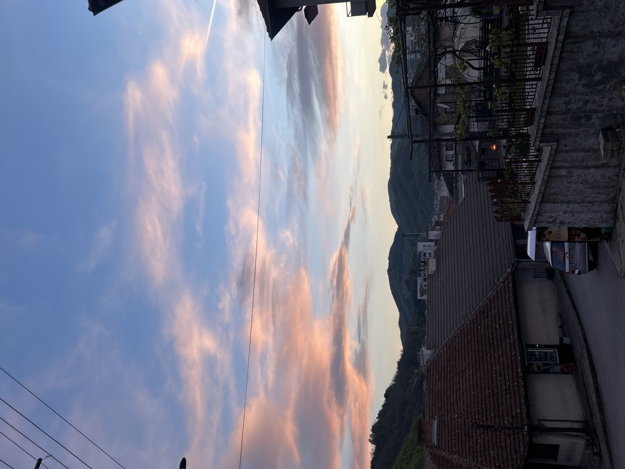 A street view in Zlatograd, Bulgaria at dusk reveals a landscape where the built environment meets natural topography. The sky is dominated by striated clouds in shades of blue and peach, possibly reflecting a sunset. The sky also shows a contrail left by a passing aircraft. Power lines cut across the lower left foreground of the sky.
On the right side of the image, a row of buildings line the street. A stone wall and ornate iron railings define the edge of one property, hosting potted plants and climbing vines. The roofs of buildings are visible, some tiled in red clay, adding texture and color to the urban landscape. A silver van is parked along the road.
In the background, mountains rise, covered in a mix of vegetation. The urban area seems nestled into the valley floor, creating a contrast between the built structures and the natural setting. A single red light is visible within the building structures.