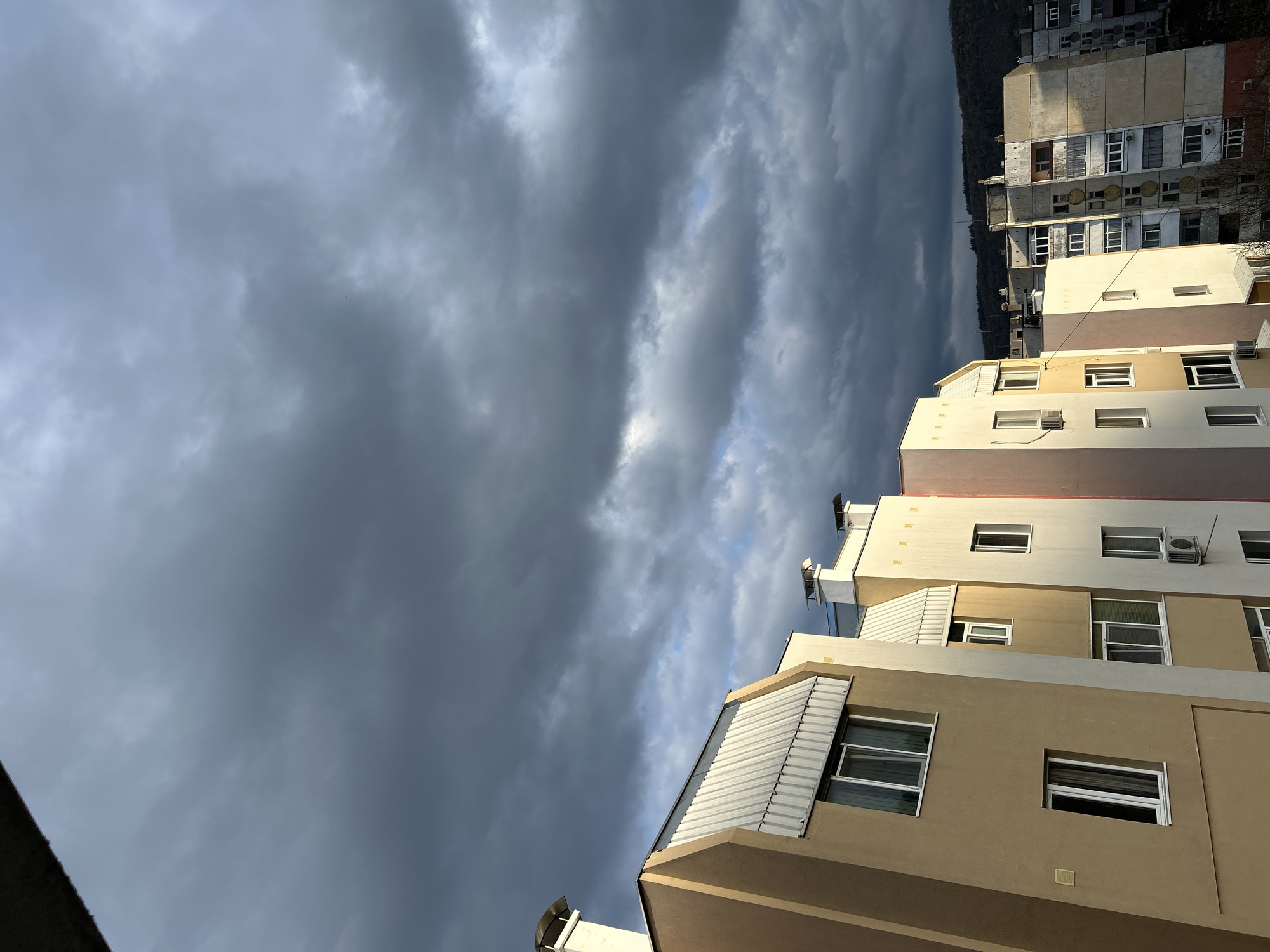 The image shows a view upward toward the sky, with buildings on the right side of the frame. The sky is filled with dark, heavy clouds. The buildings are several stories tall, with a mix of beige and white exteriors. Windows and air conditioning units are visible on the facades. In the far background, more buildings and trees are discernible. Based on the location clues provided, this scene depicts an urban landscape in Shumen, Bulgaria.
