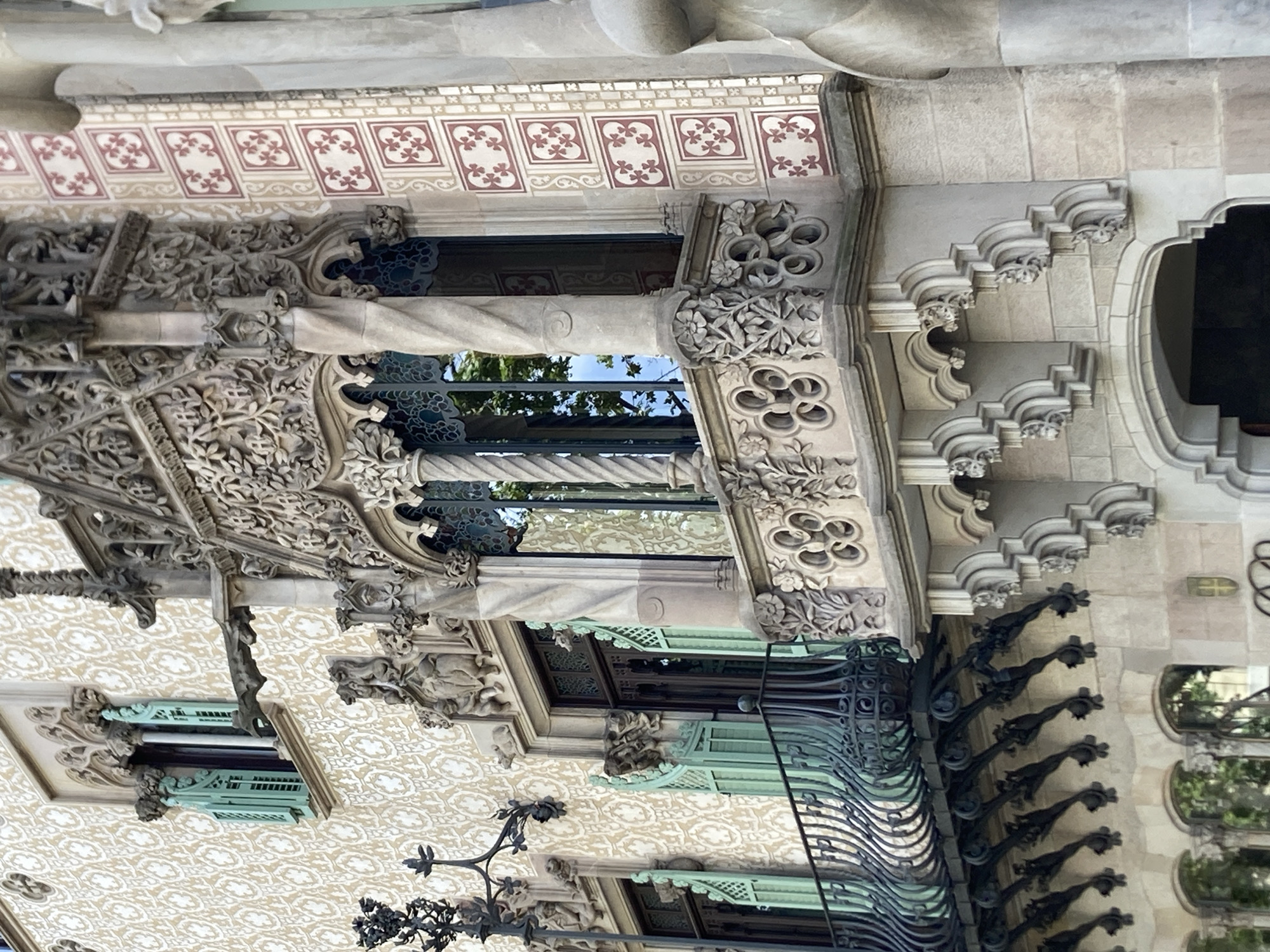 This is a detail shot of the facade of Casa Batlló, located on Passeig de Gràcia in Barcelona, Spain. The image focuses on a section of the building's upper floors, showcasing its distinctive modernist architecture.
The primary subject is an ornate balcony with intricate stone carvings and a decorative wrought-iron railing. The balcony juts out from the building, supported by a series of sculpted brackets that resemble gargoyles. Above the balcony, a large window is framed by elaborate, organic-looking stonework, with twisted columns on either side. The window itself has dark metal mullions and reflects the sky and trees.
To the left of the balcony, another window is visible with light green shutters, partially open, revealing dark wood. This window is set into a wall covered in a textured, light-colored pattern, possibly ceramic tiles or stucco. Above this window, a grotesque-like figure, likely a decorative drainpipe, extends outwards.
To the right of the main balcony, a section of the wall is adorned with a repeating pattern of square tiles, each containing a red floral motif on a light background. Further to the right, a large, smooth, grey column is partially visible, hinting at the building's overall structure.
The overall impression is one of rich detail, organic forms, and a high level of craftsmanship, characteristic of Antoni Gaudí's architectural style. The lighting suggests it is daytime, with natural light illuminating the facade. There are no people or specific actions depicted in this focused view of the architecture.