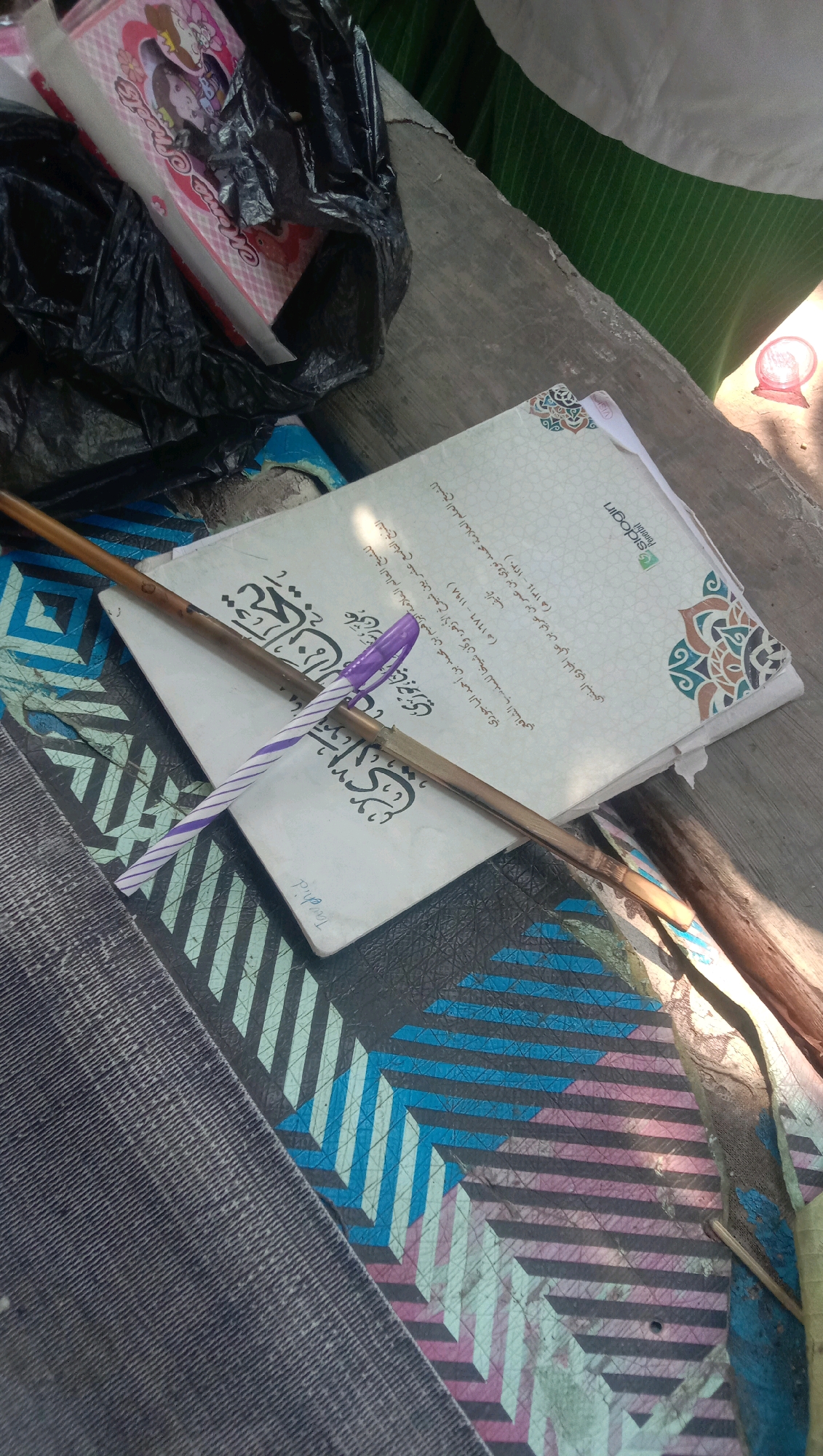 The image depicts a close-up, slightly overhead view of a table with several objects. In the foreground, a wooden stick lies horizontally across two books. One book, on top, is white with Arabic script and decorative borders. A purple and white striped pen is placed on top of this book, resting on the stick. The second book, underneath, also has Arabic script. A black plastic bag is visible in the upper left corner, containing a pink and white patterned item, possibly a box of tissues. The table surface appears to be made of wood. In the background, a green and white striped fabric is visible, along with a hint of a person's arm and shoulder. The lighting suggests an outdoor setting, possibly in a shaded area. The image seems to be taken in the city of Ngempit, Indonesia, based on the provided context.