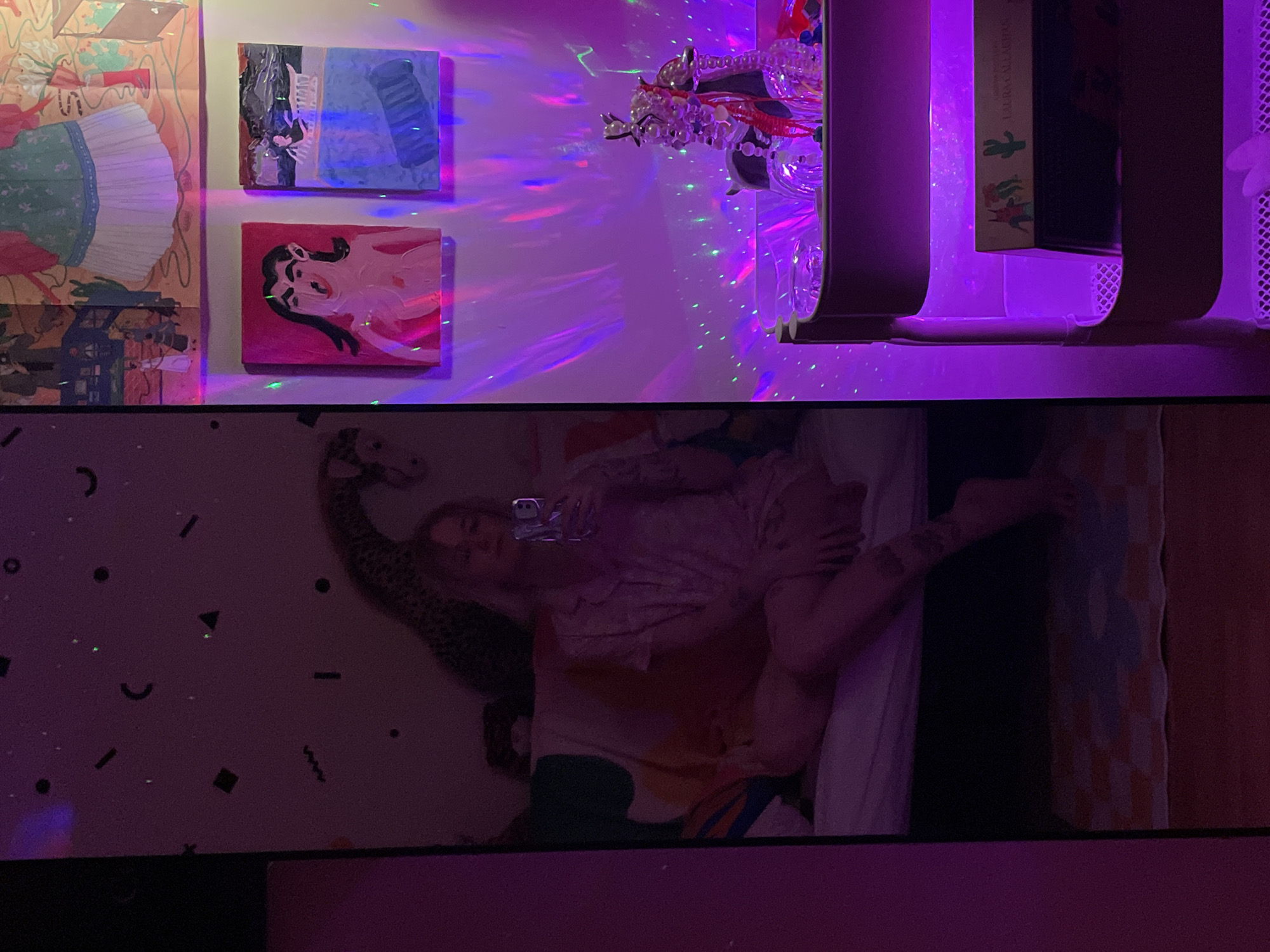 The image depicts a room interior, likely a bedroom, under purple and blue lighting. On the upper wall, there are several pieces of art. From left to right: a colorful artwork featuring a figure in a dress, a blue abstract painting, and a red-toned painting of a figure. There is a clear decorative object with bead strands and a shelf with books.
Below, visible in a reflective surface, is a person reclining on a bed. They are holding a phone, possibly taking a picture. The person is wearing a patterned shirt and is resting against a leopard print pillow. The bedding includes a white sheet and colorful patterned blankets. There are scattered geometric patterns on the wall behind the bed. The location information, Belgrade, Serbia, is not definitively indicated.