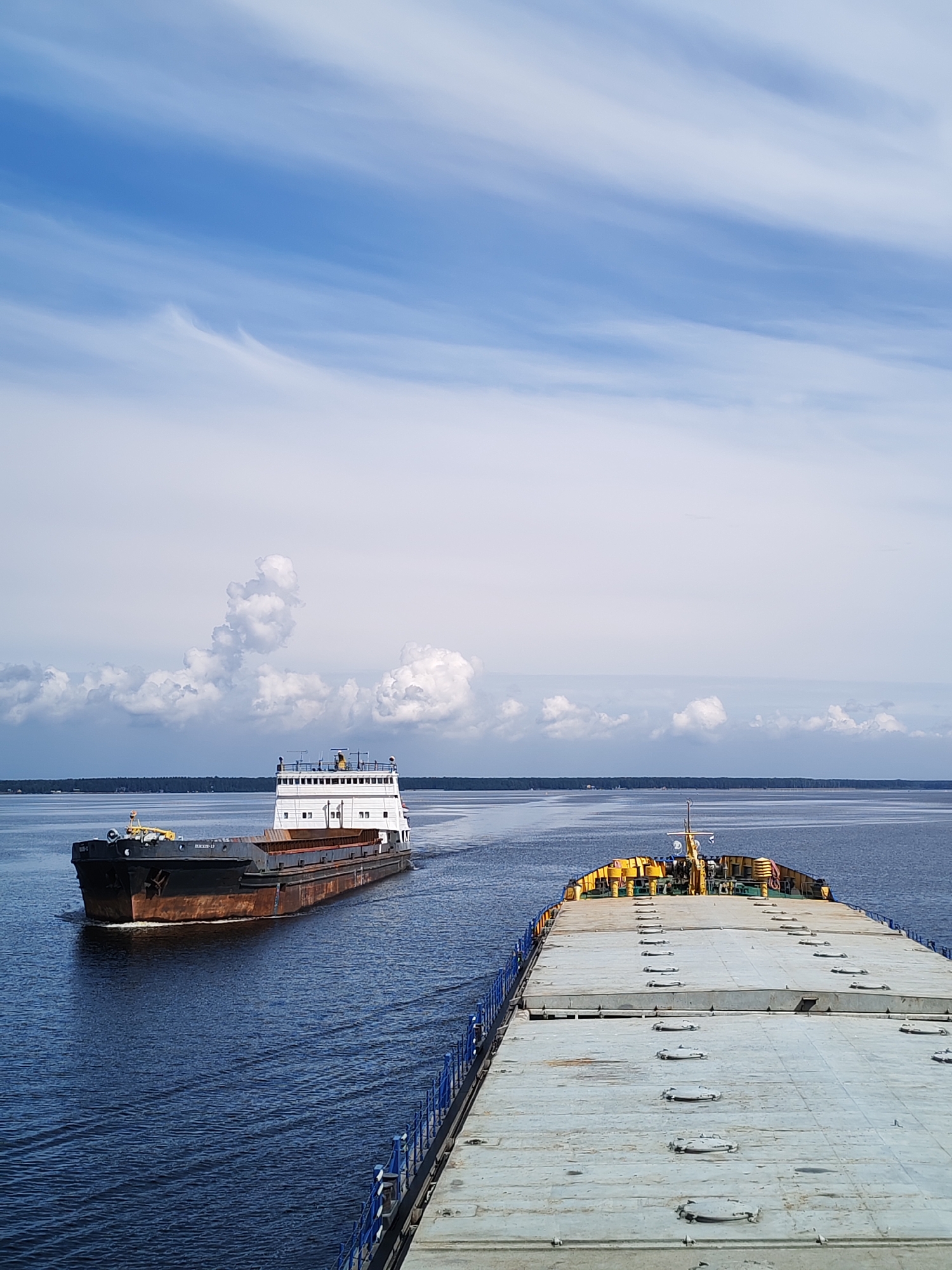The image depicts a wide, calm body of water under a partly cloudy blue sky. Two large cargo barges are visible. The barge in the foreground is positioned so that its deck, covered with numerous hatches, fills the lower right portion of the frame. A blue railing runs along the edge of this barge. The second barge is further in the distance, moving away from the viewer and towards the left side of the frame. It has a dark, rusty hull and a white superstructure. The horizon is marked by a dark, treeline representing land. The scene is one of transportation and industry on a waterway. The location is identified as the city of Kostyaevka, Russia.