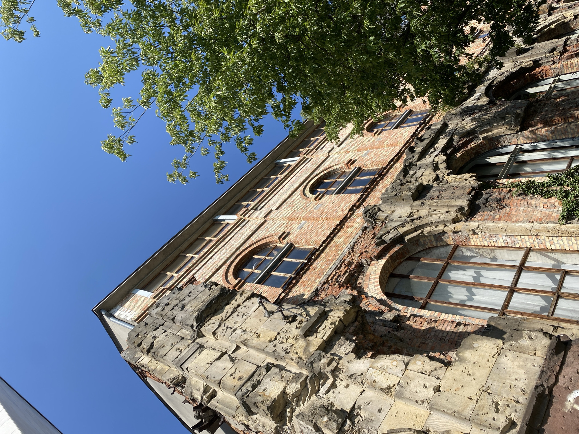This image shows the exterior of a partially damaged brick building under a clear blue sky, with green foliage from a tree in the foreground on the right. The building appears to be an old structure, with a section of its stone facade severely weathered and pitted, suggesting it may be a historical site or a building that has sustained damage. The brickwork is exposed in places, revealing the age and construction of the building. There are several tall, arched windows with wooden frames and white curtains visible. The building's architecture suggests it is located in an urban environment. Specifically, this building is located at the intersection of Ulica Marszałkowska and Aleje Jerozolimskie in Warsaw, Poland, and is known as the "Prudential" building, a landmark that survived the Warsaw Uprising.
