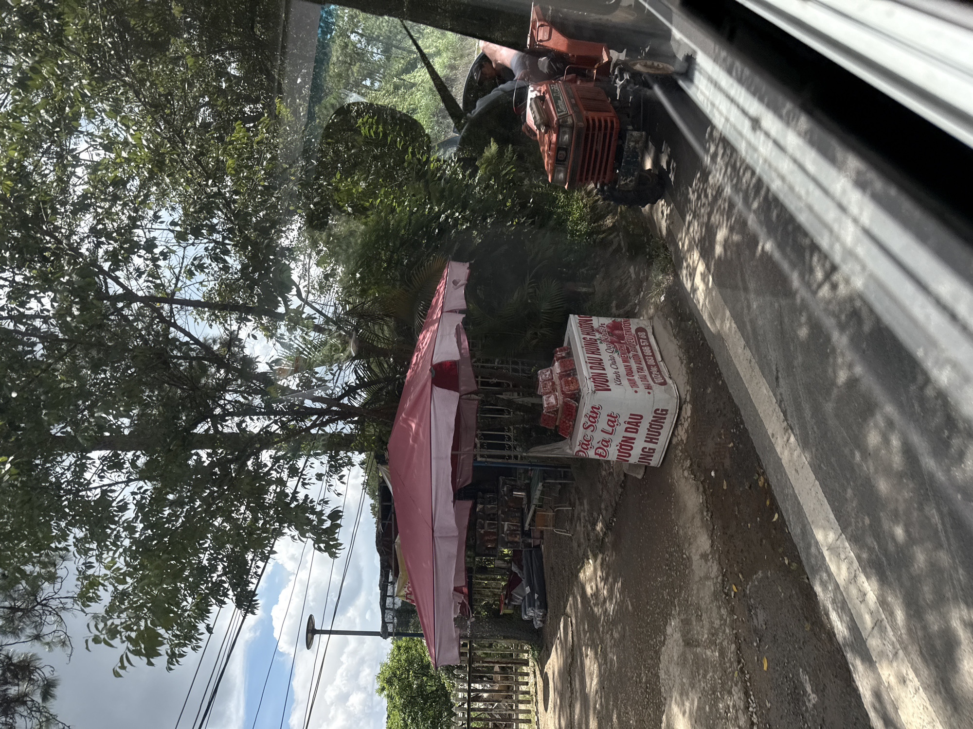 In Thôn 6, Vietnam, a roadside stall with a pink and white striped awning displays various goods, including boxes of what appear to be strawberries and other packaged items. Behind the stall, stacked rolls of dark material are visible. A red tractor, driven by a person wearing a hat, is parked to the right of the stall on the dirt road. The scene is bordered by a wooden fence and lush green foliage, with a cloudy sky overhead. A street lamp is also visible in the background. The image appears to be taken from inside a vehicle, as evidenced by the reflections on the glass.